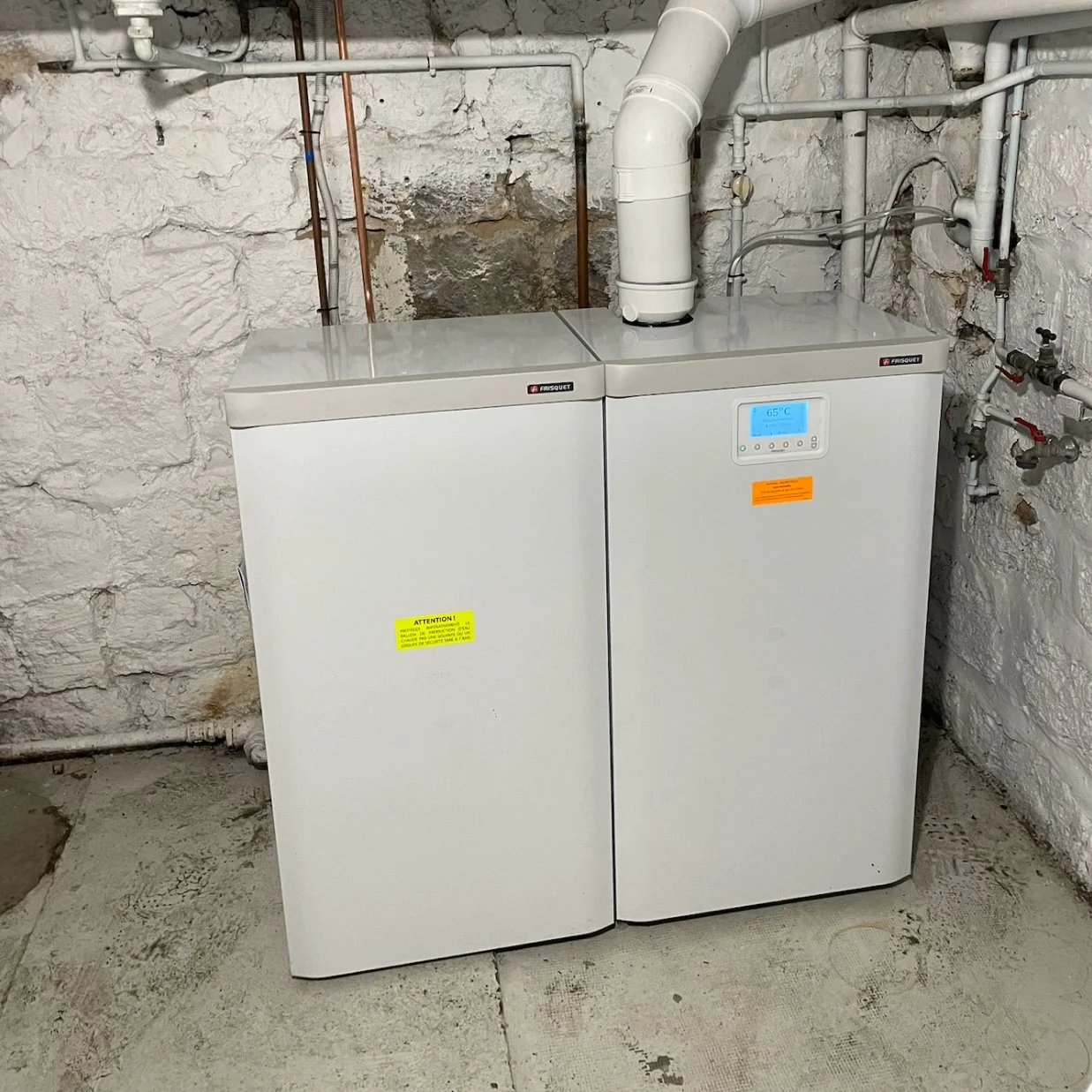 Chauffe-eau ou chaudière dans une pièce avec murs en pierre, affichant une température de 65°C sur un écran numérique, connecté à des tuyaux en PVC et en cuivre.