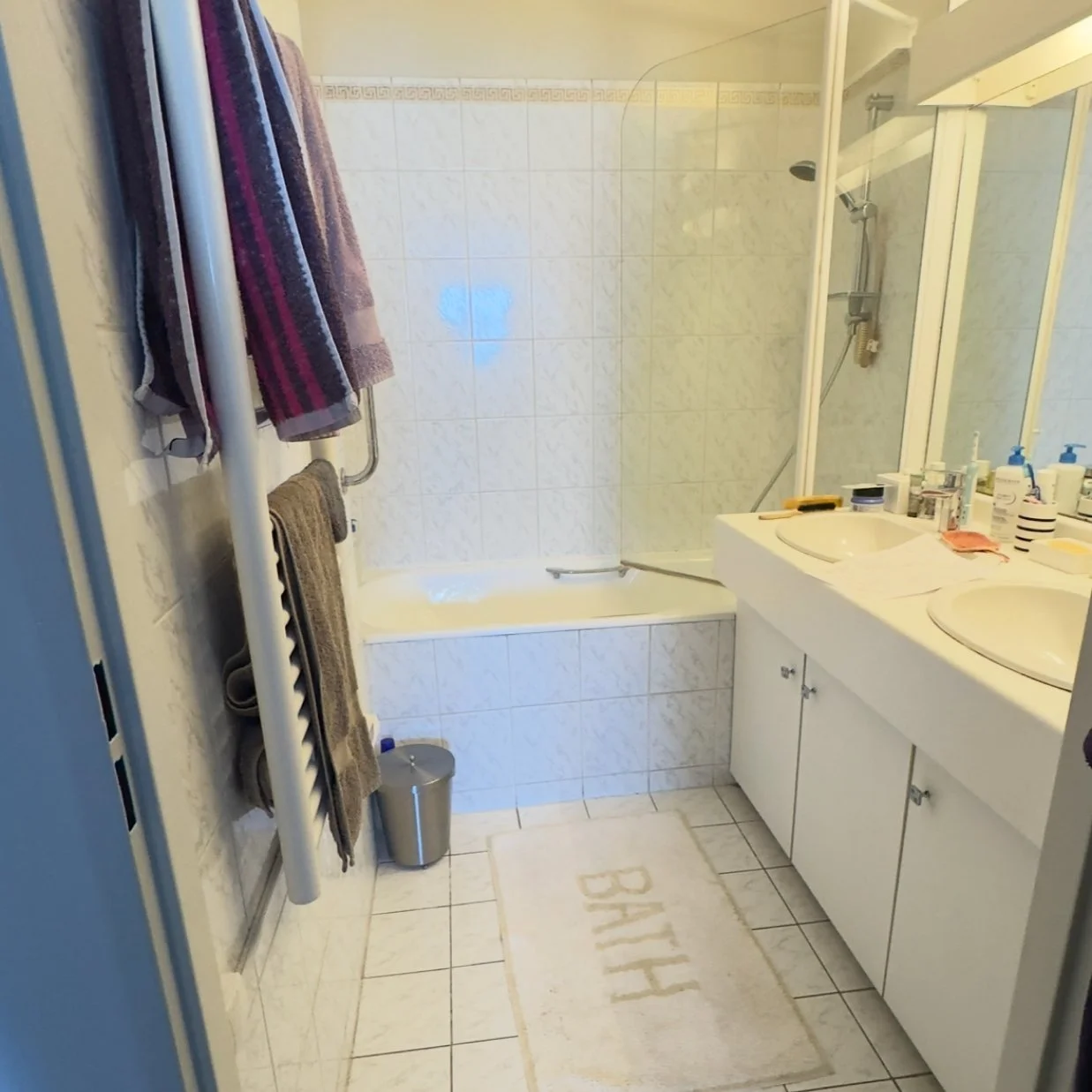 Salle de bain avec baignoire, double lavabo sur meuble blanc, miroir, étagère avec divers produits, serviettes suspendues, tapis de bain avec le mot 'BATH', porte de douche en verre.
