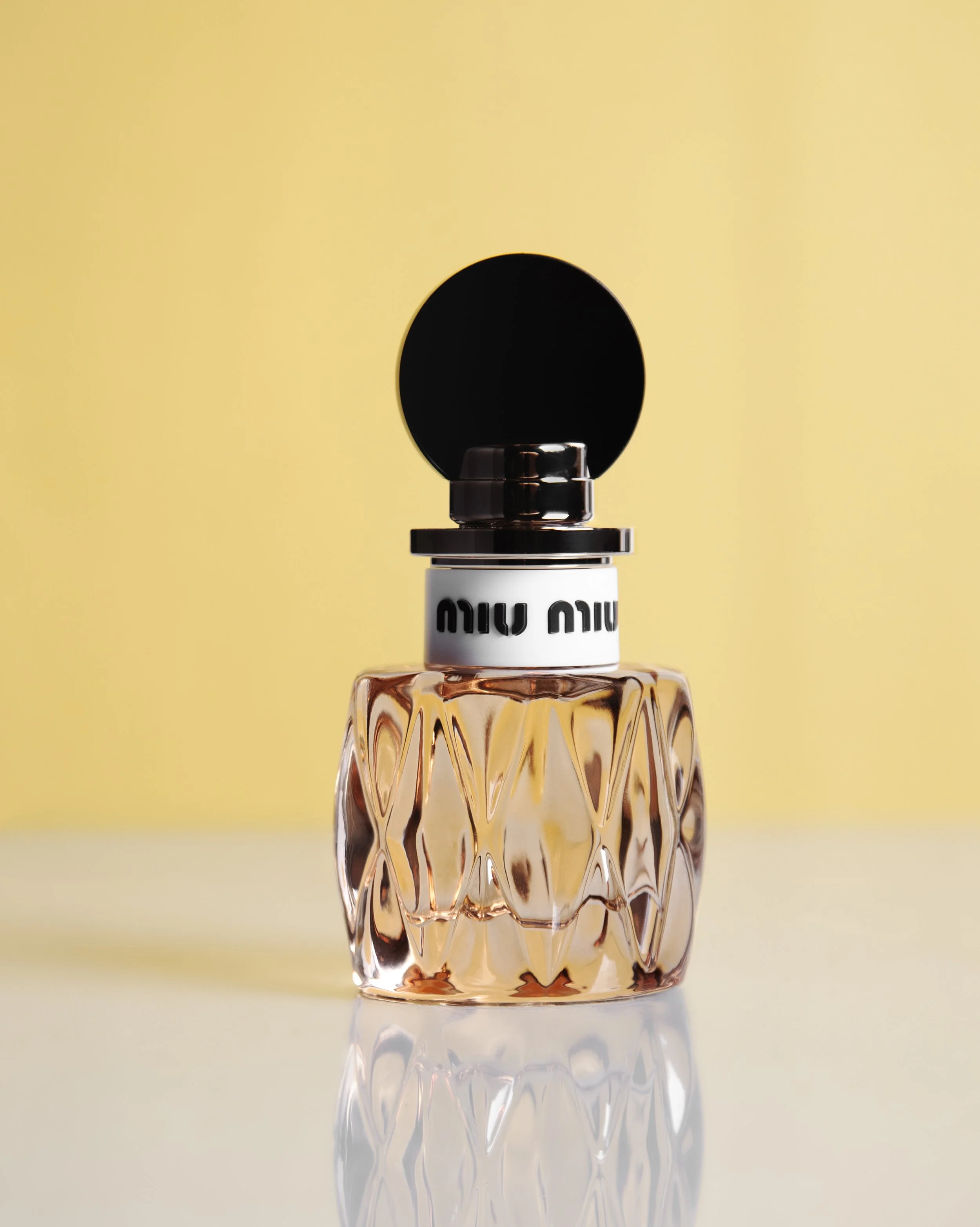 A perfume bottle with a textured glass design, a black round cap, and a white label with black text, set against a yellow background.