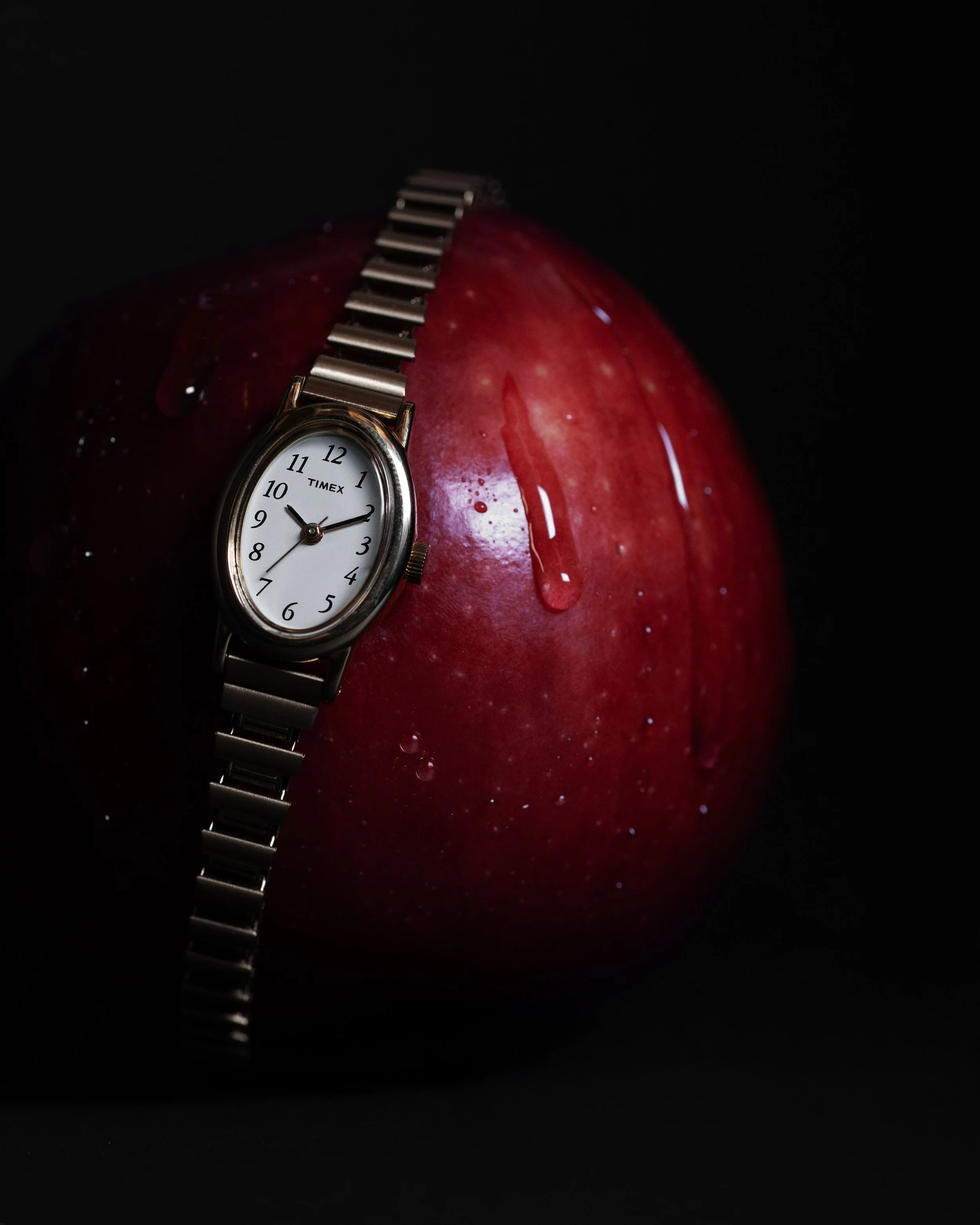 A golden wristwatch with a white face and black numbers, displaying the time 9:10, is placed on a shiny red apple with water droplets on its surface, against a black background.