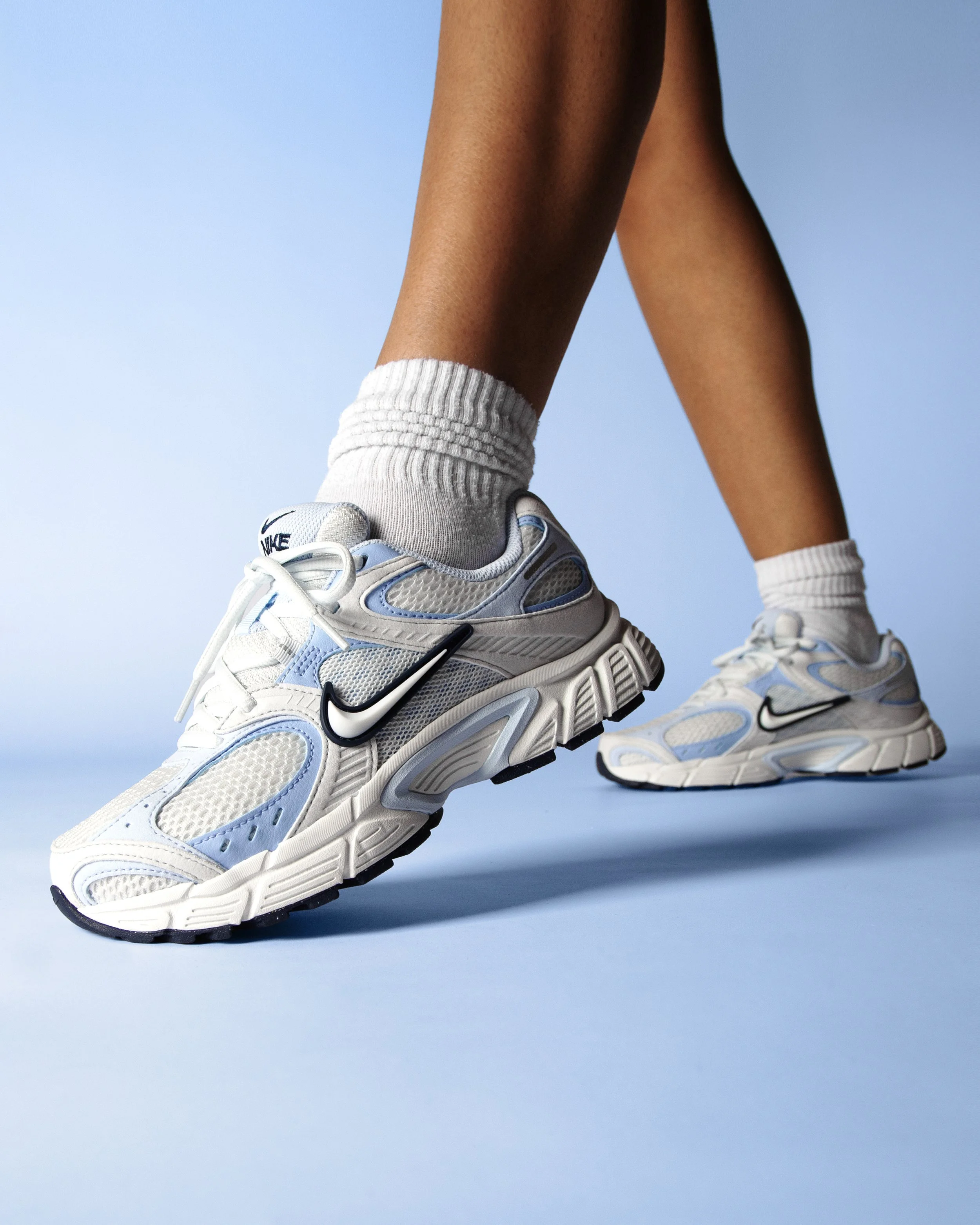 Close-up of a person wearing white and blue Nike running shoes, white socks, and standing against a light blue background.