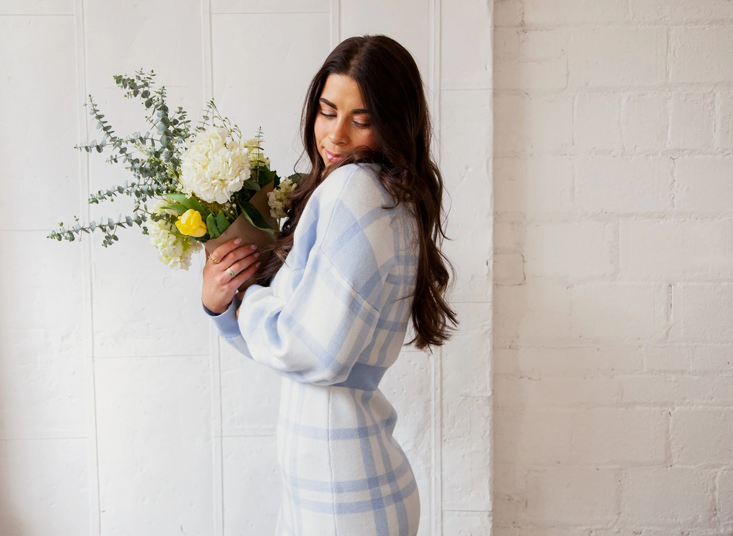 A woman with long dark hair holding a bouquet of white and yellow flowers, standing against a white brick wall, dressed in a light blue and white checkered shirt.