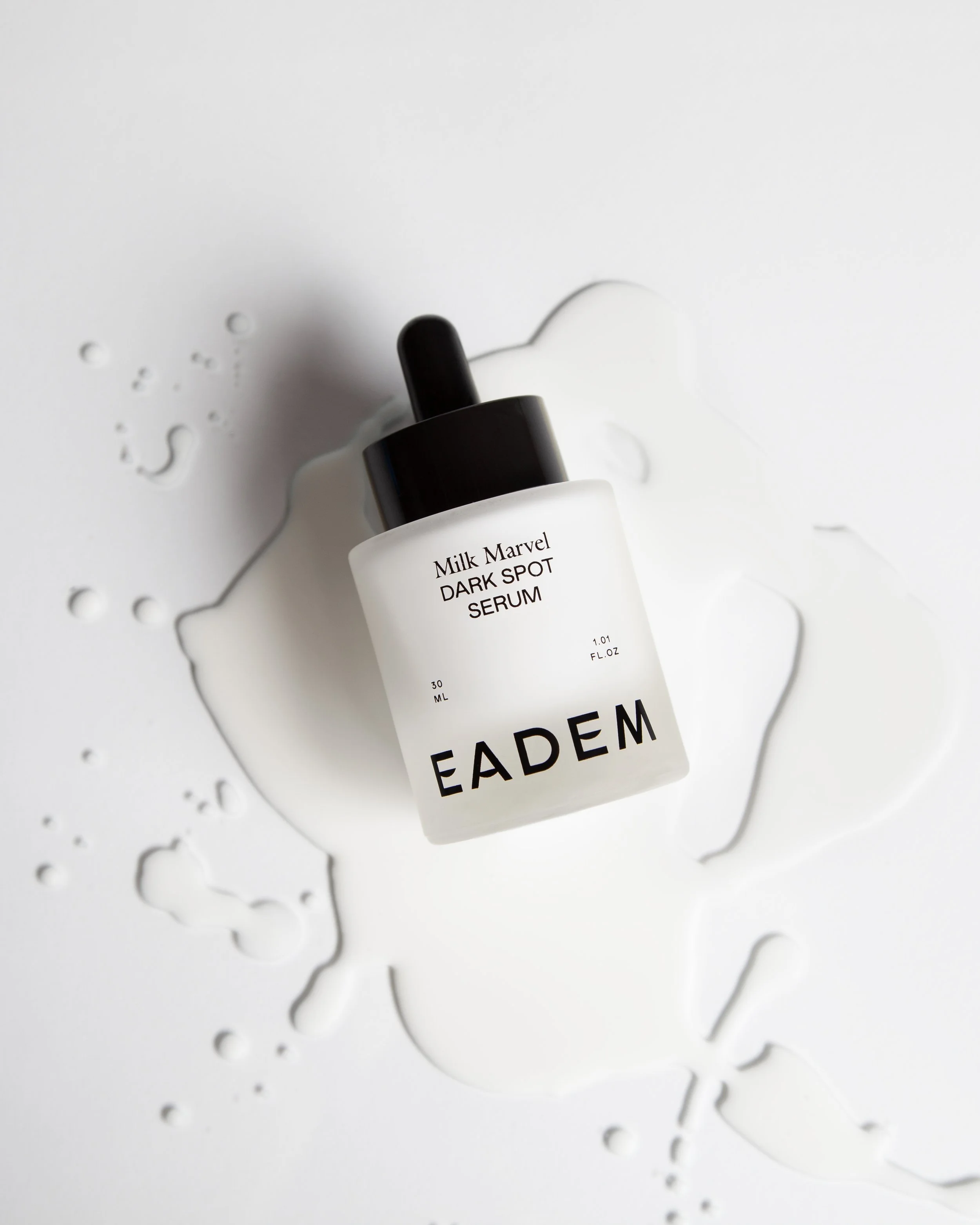 A white dropper bottle labeled 'Milk Marvel Dark Spot Serum' with spilled serum droplet on a white surface.
