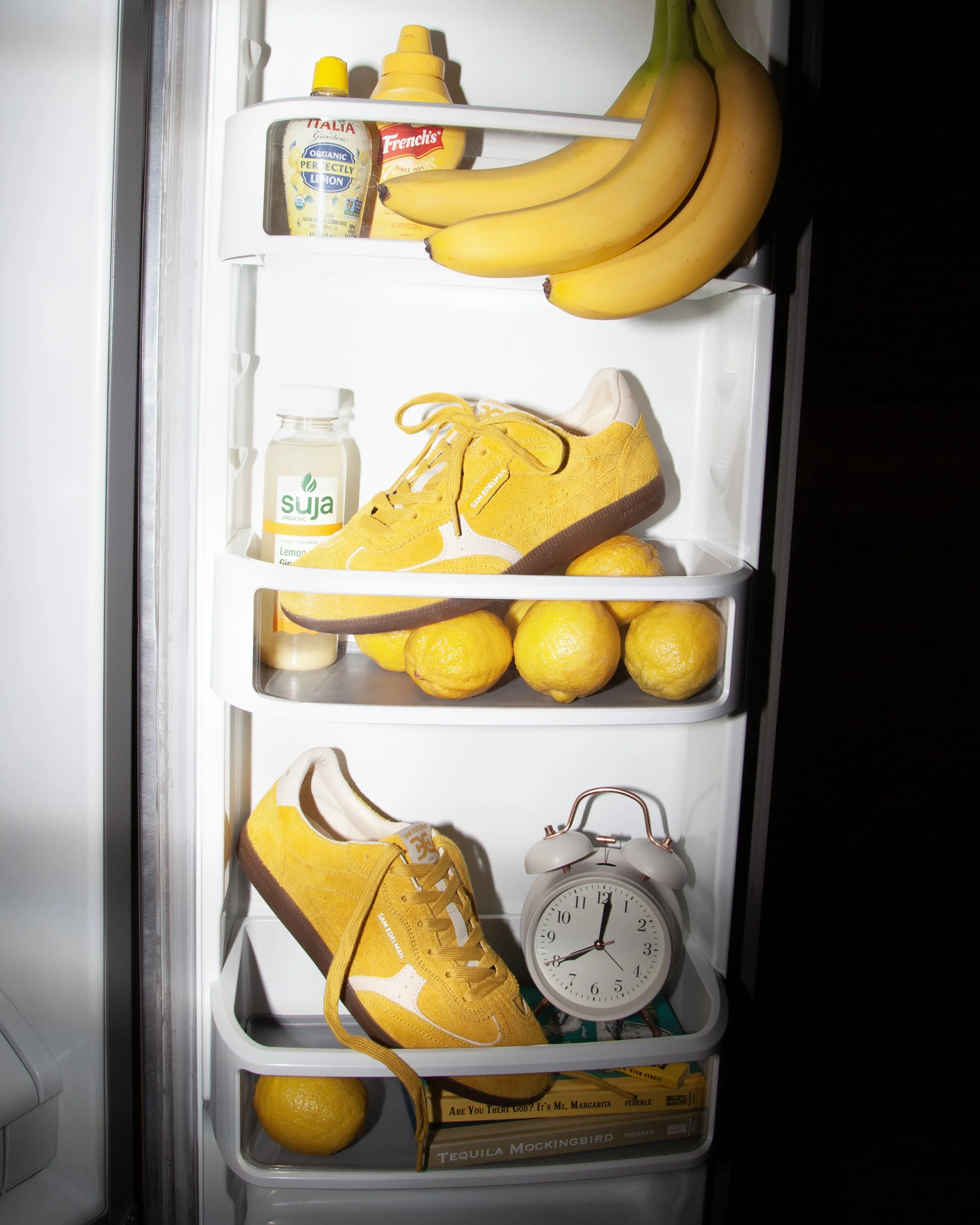 Open refrigerator door with a bunch of bananas hanging on the door, a yellow sneaker on a shelf, a lemon, a bottle of Suja organic lemon ginger juice, a container of French's mustard, a container of Italy Garden organic lemon slices, a vintage alarm 