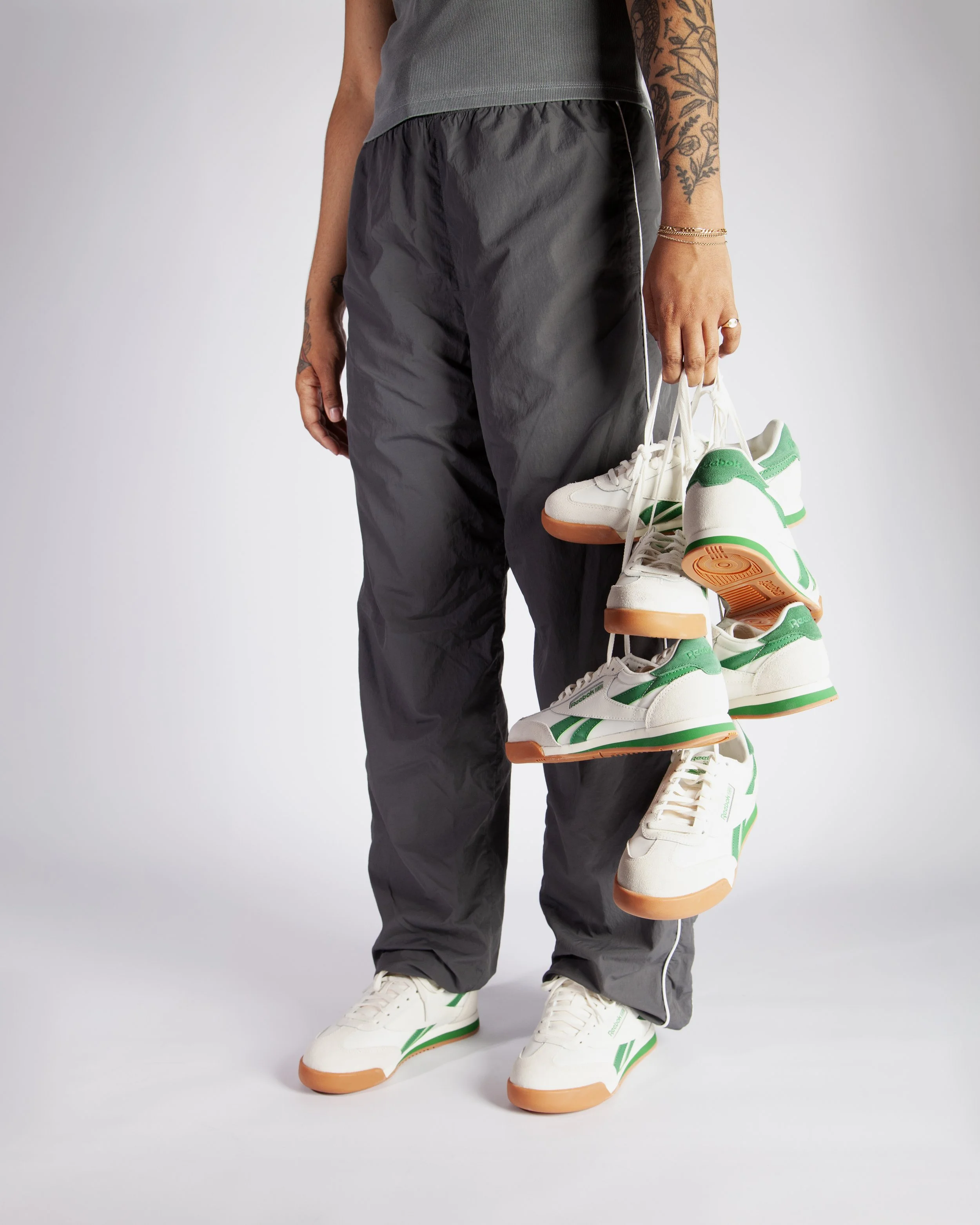 Person dressed in gray athletic pants and a gray top, holding a pair of white sneakers with green accents, against a plain background.