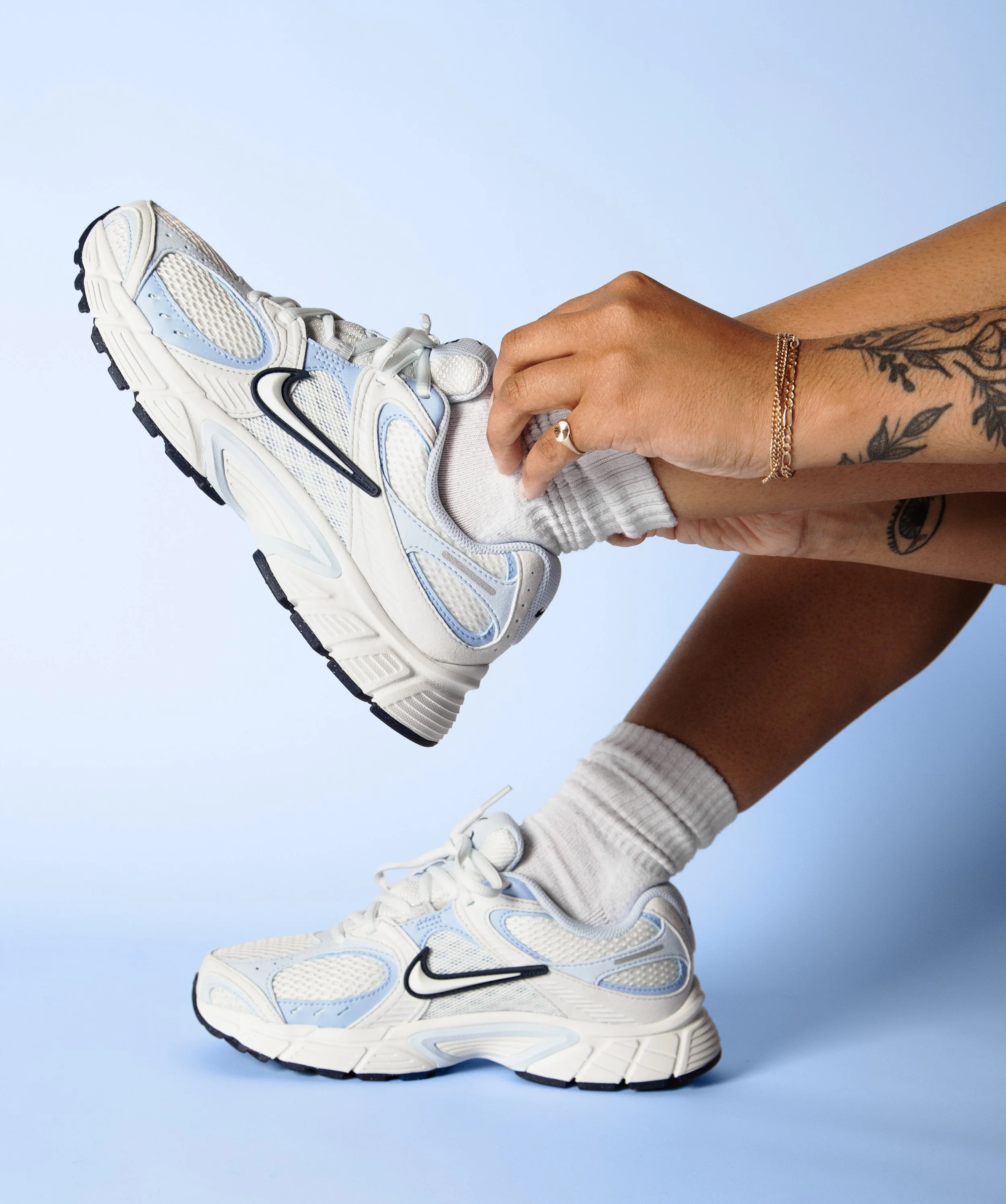 Person wearing white sneakers with blue and black accents, white socks, and shorts, stretching against a light blue background. The person has a tattoo on their forearm and wears a gold bracelet and a ring.