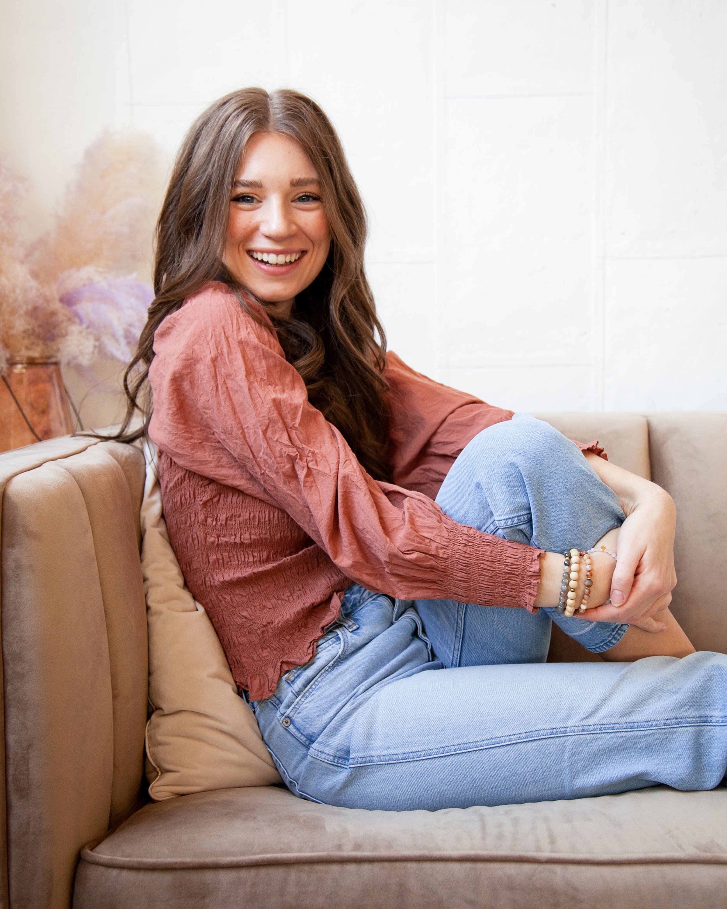 A young woman with long wavy brown hair smiling and sitting on a beige sofa, wearing a pink long-sleeve blouse and light blue jeans.