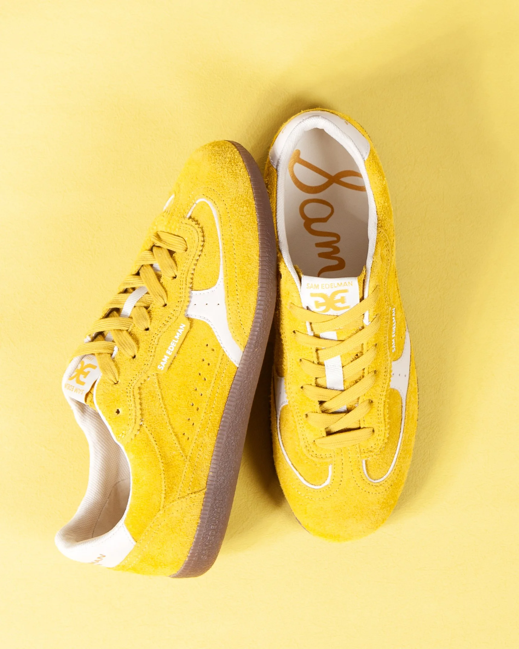 Pair of yellow sneakers with white accents, placed on a yellow background, with one shoe showing the brand label 'Sam Edelman' inside.