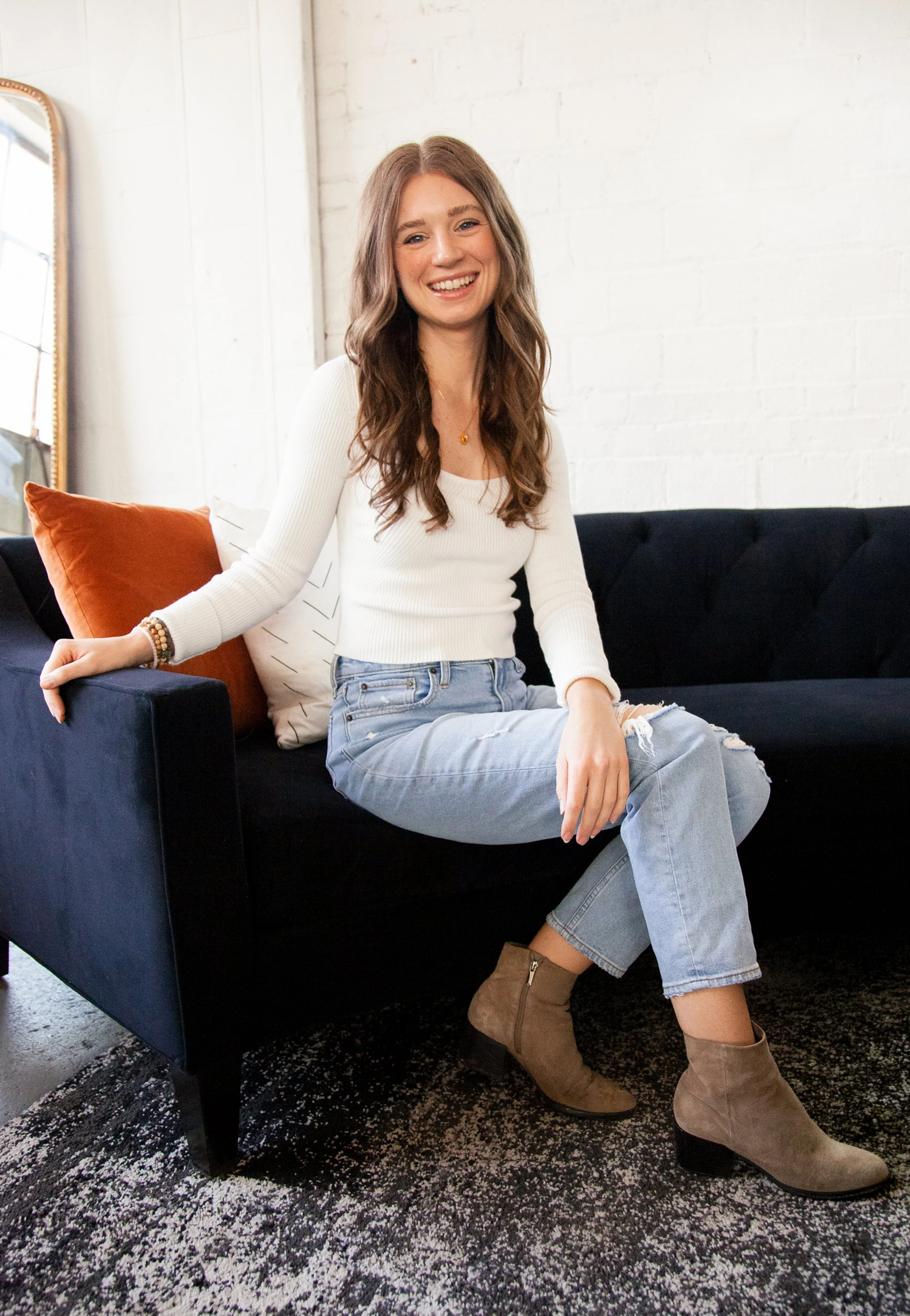 A young woman with long wavy brown hair sitting on a dark blue sofa, smiling, wearing a white long-sleeve top, ripped jeans, and tan suede boots in a modern indoor setting.