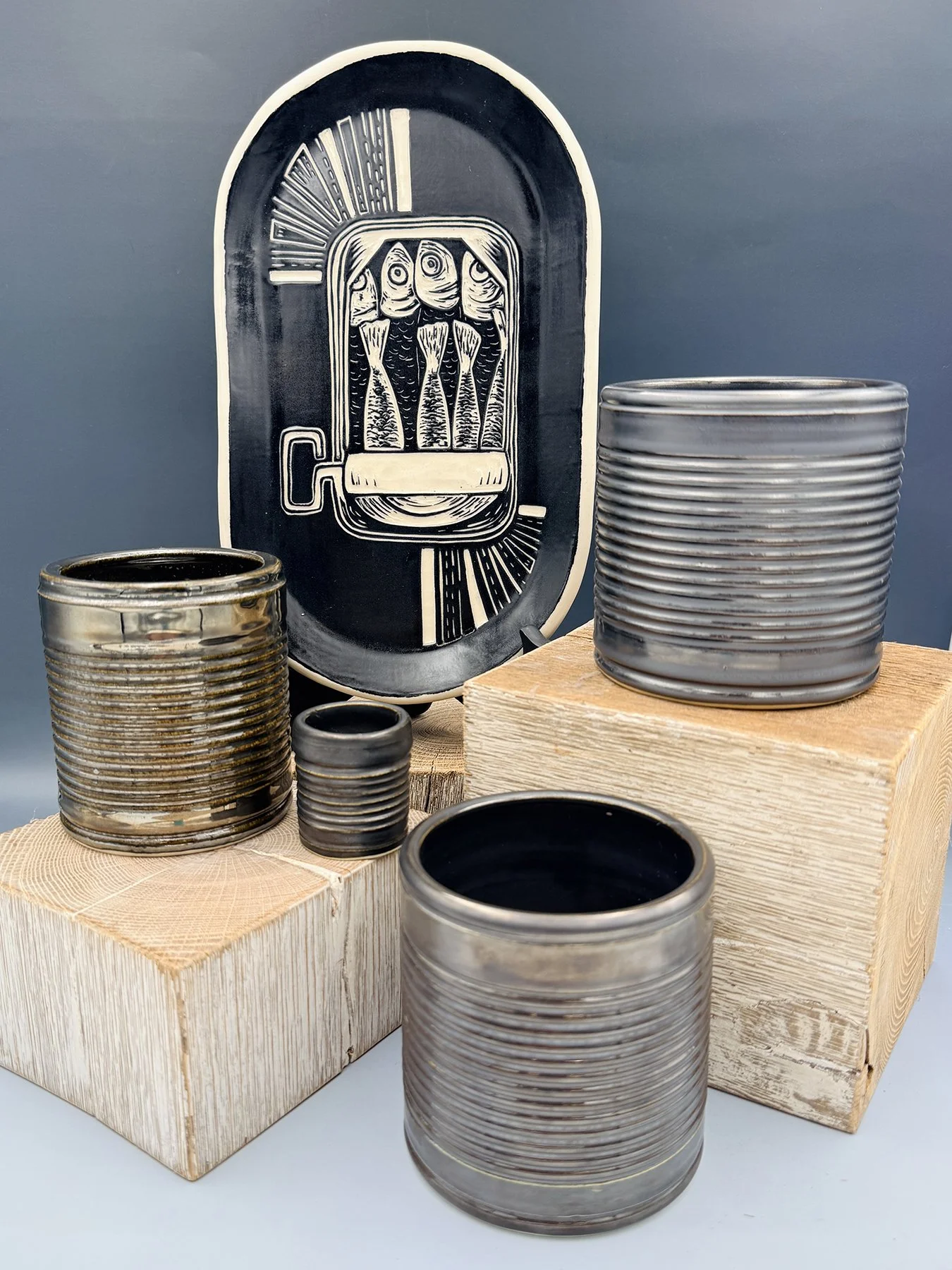 Handmade ceramic collection in dark metallic glaze — oval platter with sgraffito sardine tin illustration and four ribbed cylindrical vessels in varying sizes, displayed on wooden blocks