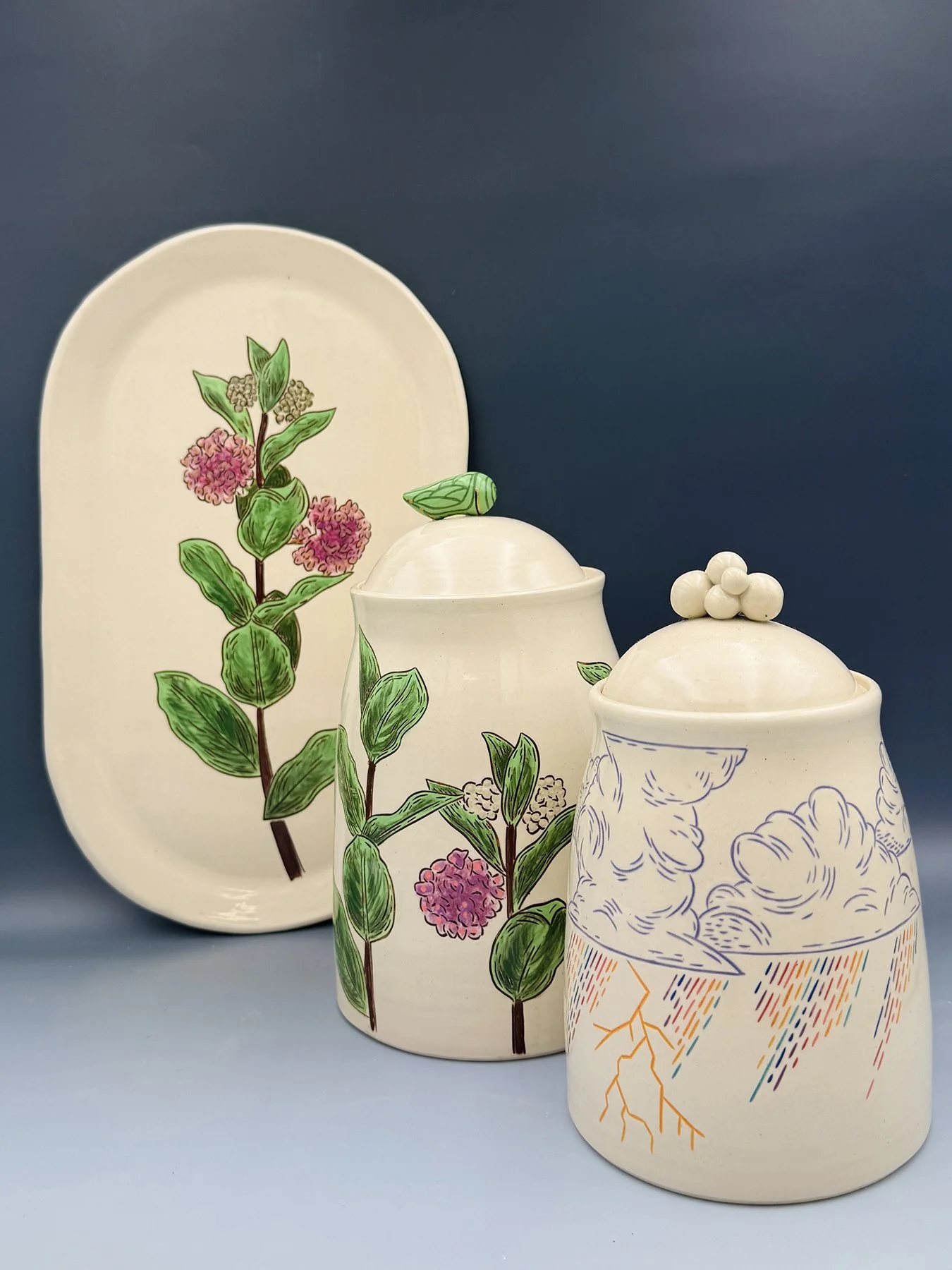 Handmade ceramic set with botanical milkweed illustration — oval platter and two lidded jars in cream glaze, one featuring a whimsical rainbow storm scene with lightning