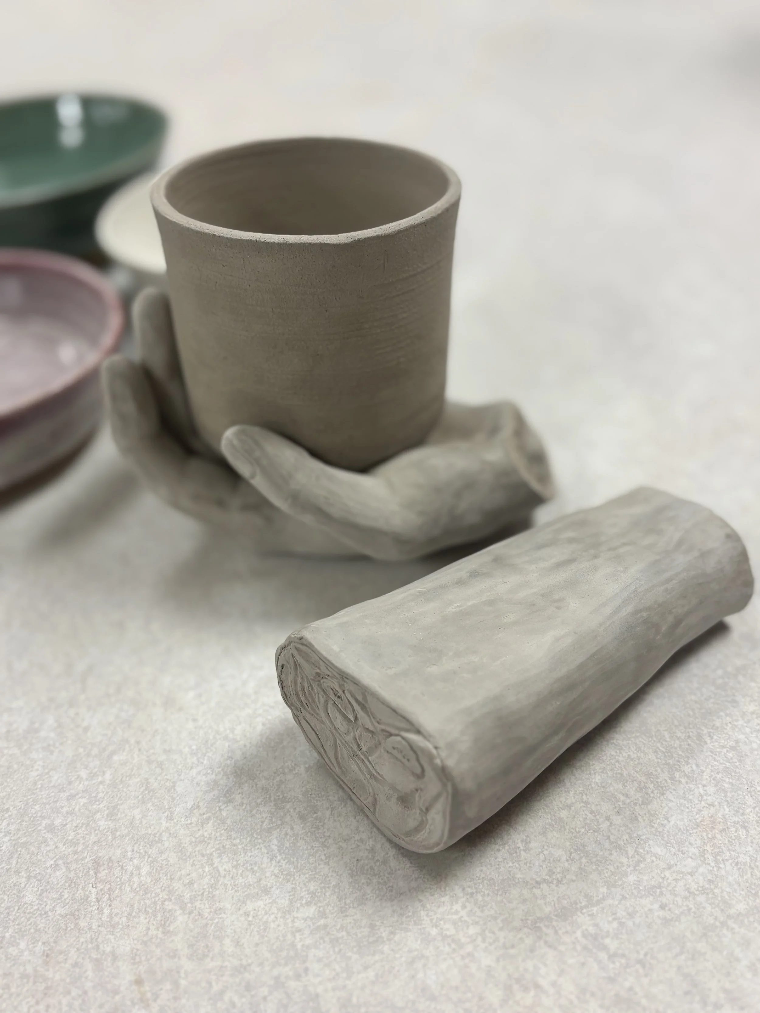 Unfired greenware wheel thrown stoneware cup cradled in a sculpted clay hand, alongside an anatomically correct ceramic arm, made by a PVCC student in Malenka Luckett's ceramics class
