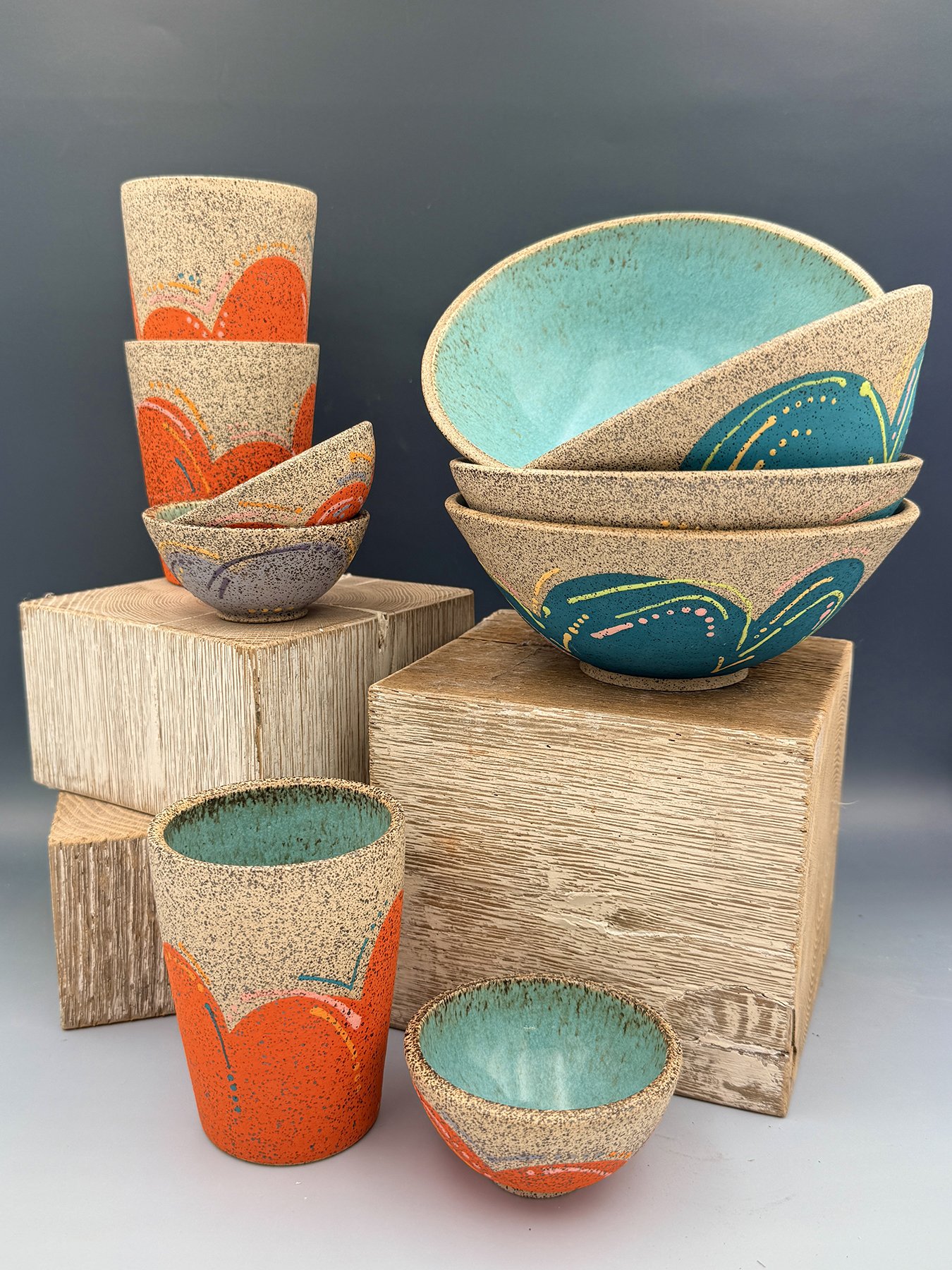 Handmade speckled stoneware bowls and tumblers with bold orange scalloped surface design, teal interiors, and colorful hand-drawn details, displayed on wooden blocks