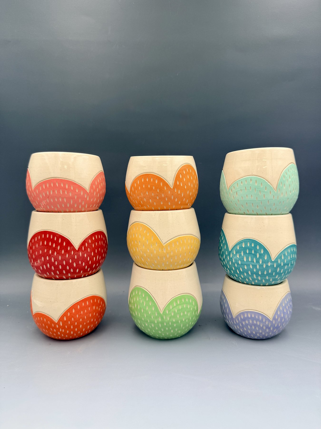 Nine handmade wheel-thrown ceramic tumblers stacked in three columns, each with a scalloped edge design in rainbow colors — pink, red, orange, yellow, green, teal, mint, and lavender