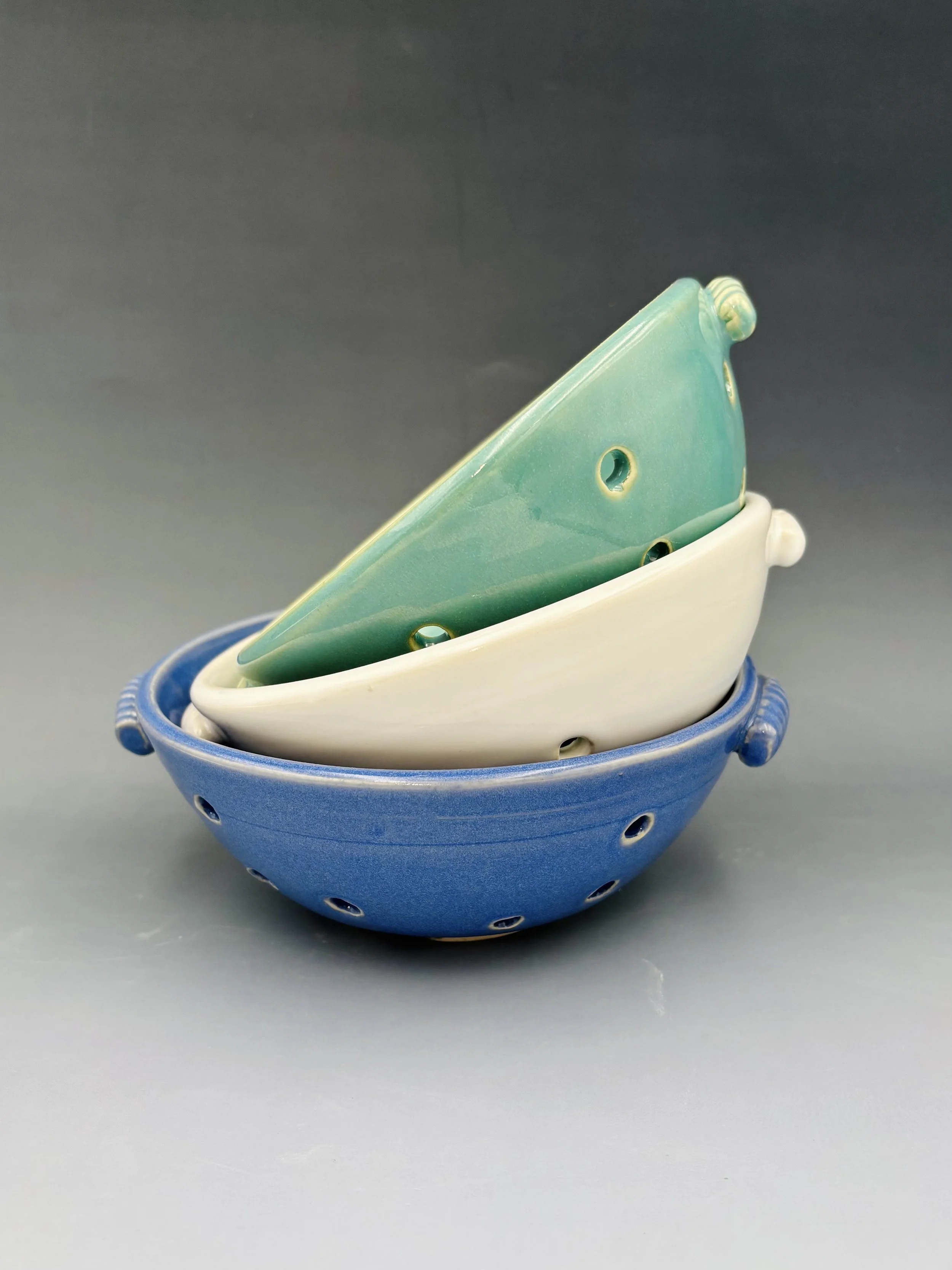 Handmade ceramic berry bowls by student Reese McCardle, stacked in three sizes in blue, white, and teal glaze with pierced drainage holes and decorative handles, made in Malenka Luckett's ceramics class