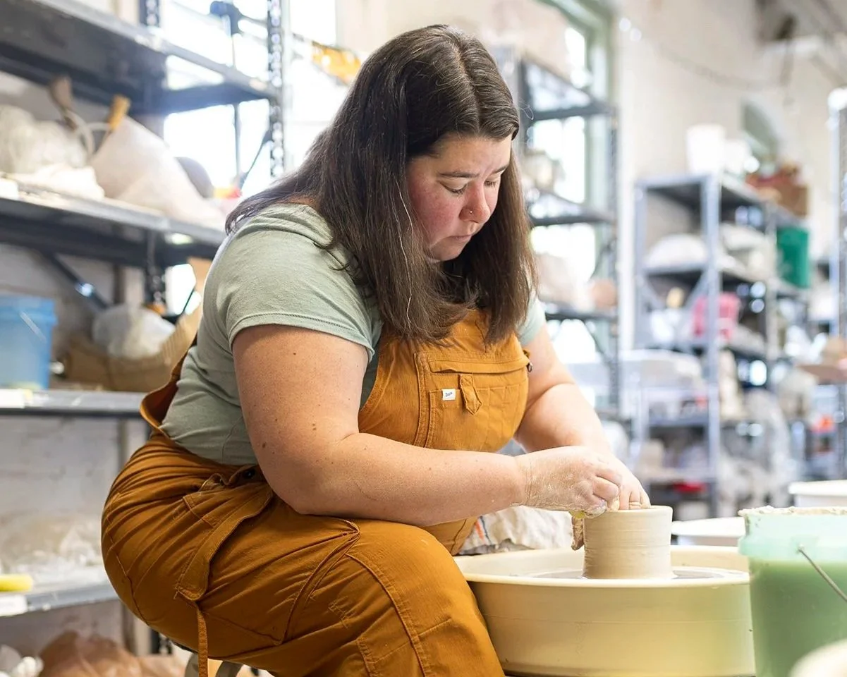 Ceramic artist Malenka Luckett seated at a pottery wheel in her studio, focused on throwing a small vessel, wearing golden overalls in a bright workshop space