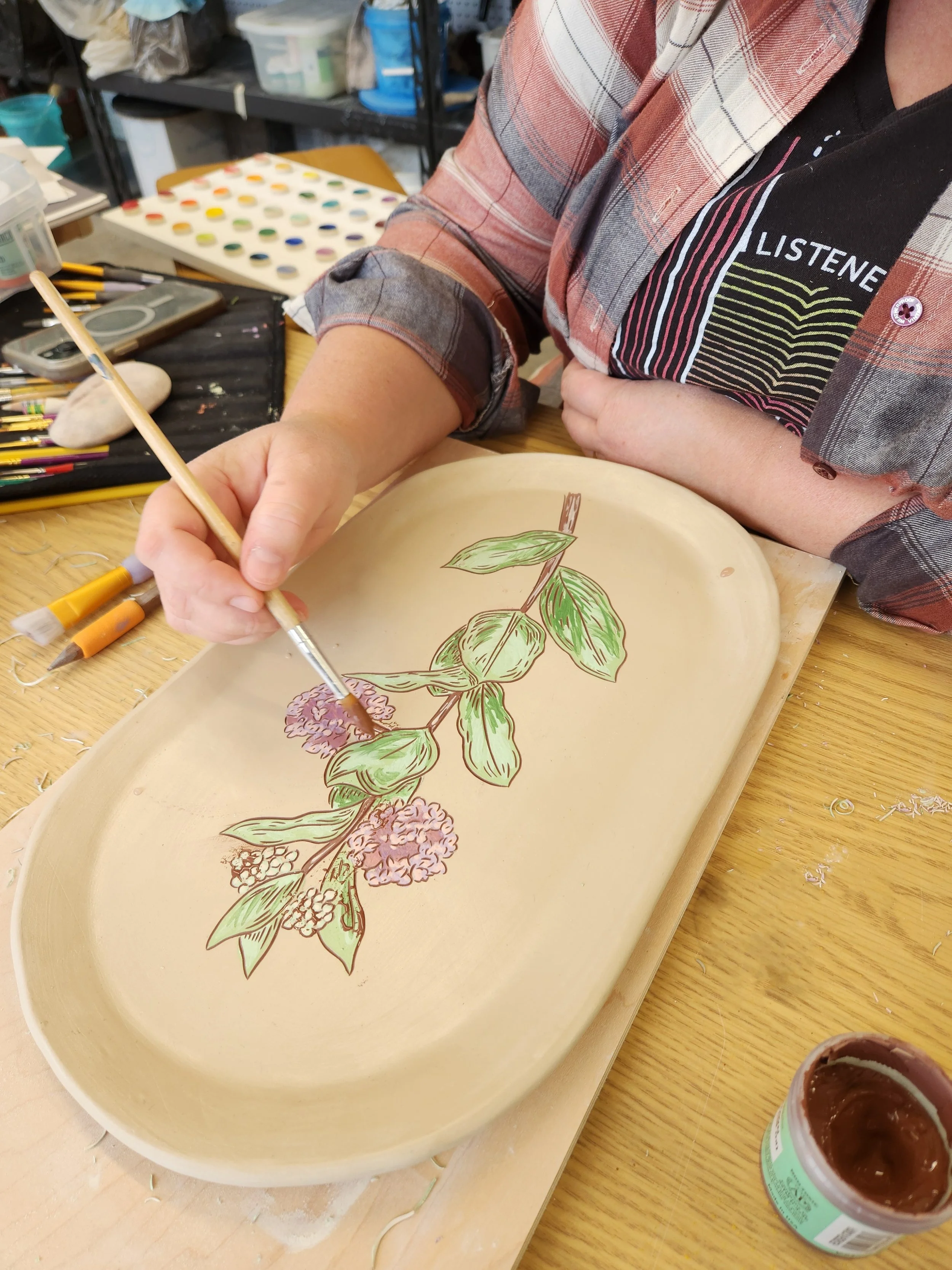 Malenka Luckett hand painting a botanical milkweed illustration onto an unfired ceramic oval platter using underglaze, with brushes and a underglaze test palette visible in the background