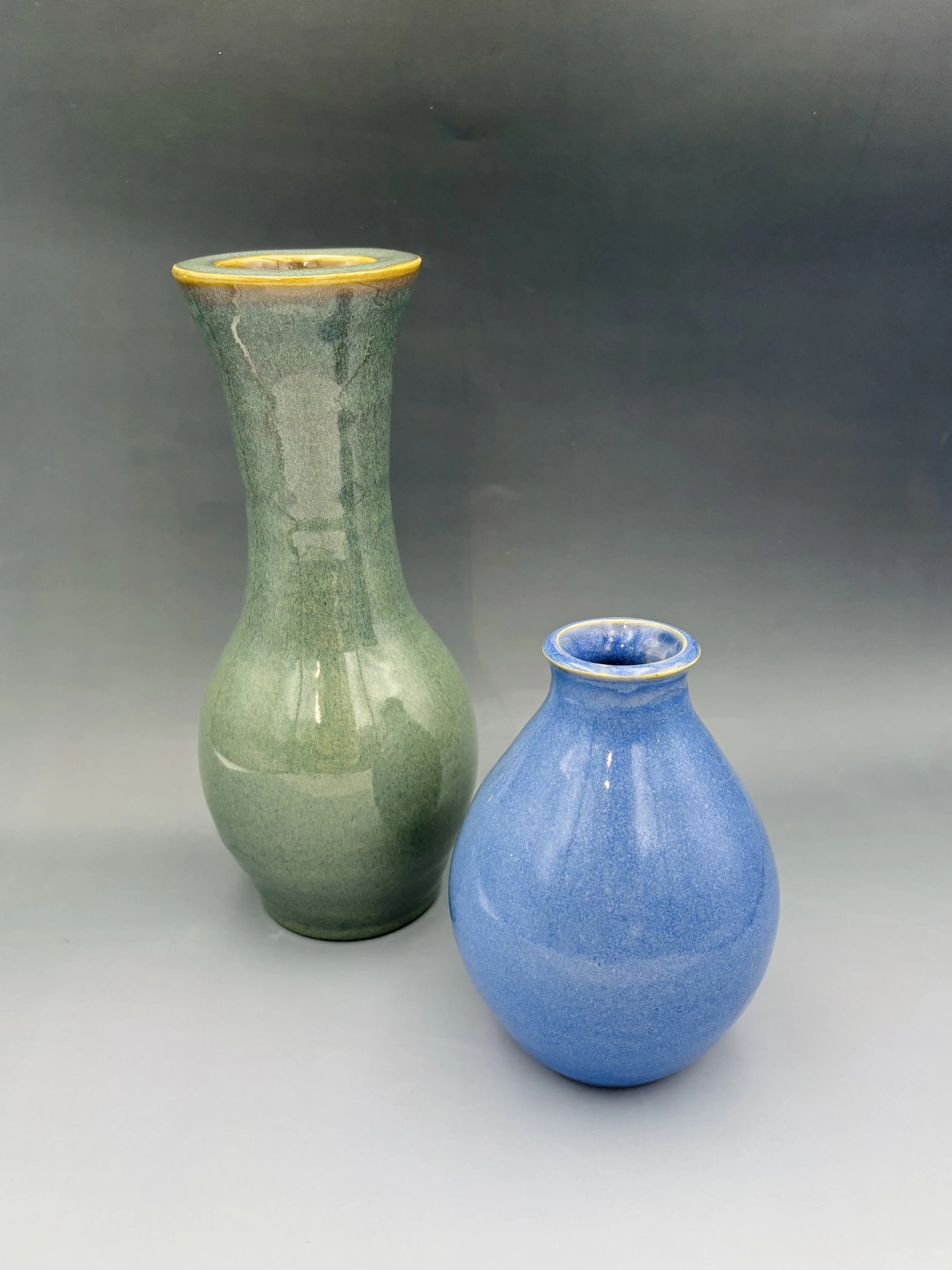 Two handmade wheel-thrown ceramic vases by student Reese McCardle — a tall green vase with yellow rim and a small round blue vase, made in Malenka Luckett's ceramics class