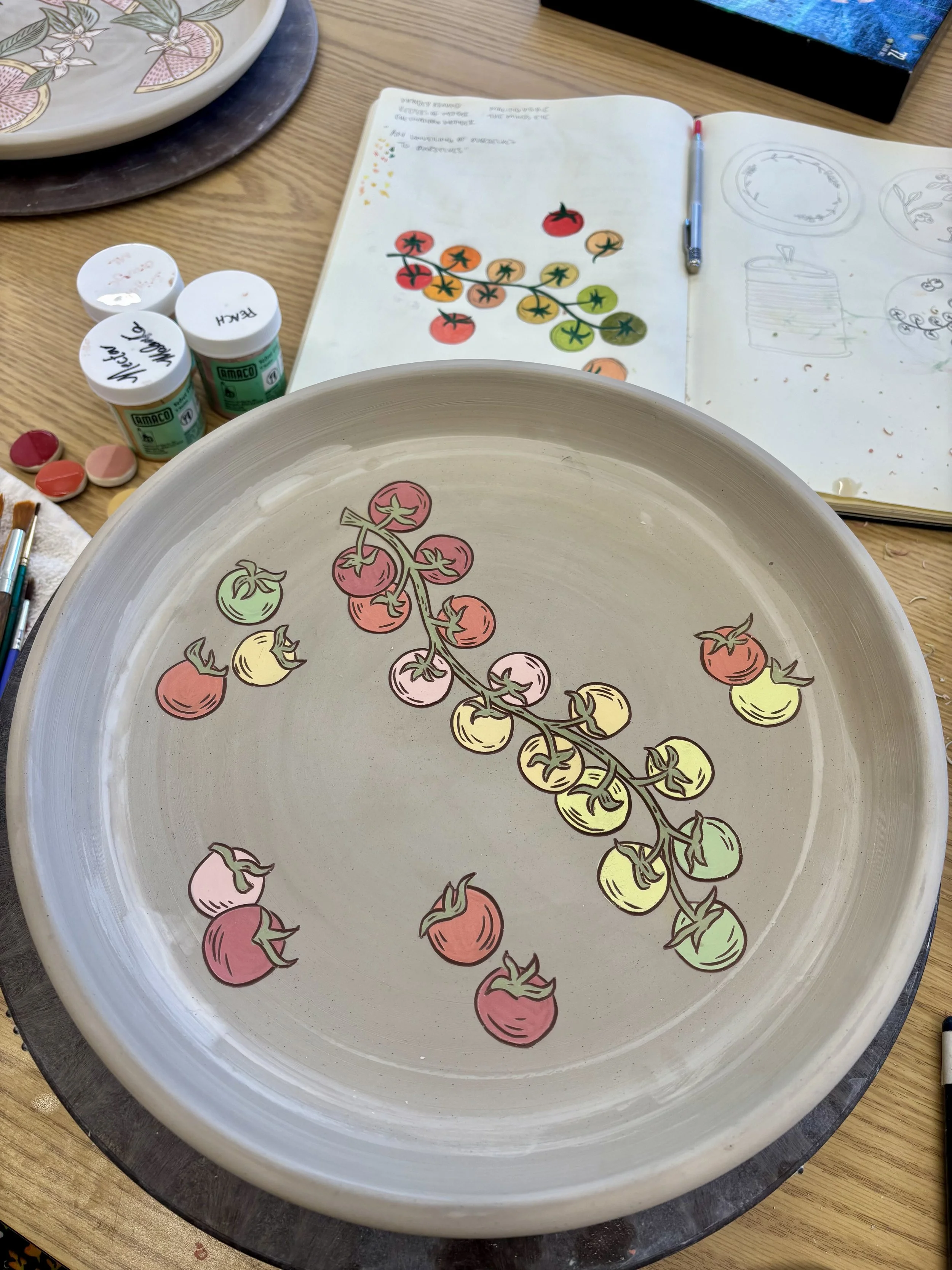 Handmade ceramic plate in progress with hand-painted heirloom cherry tomato illustration in red, pink, yellow, and green, shown alongside a sketchbook and underglaze jars