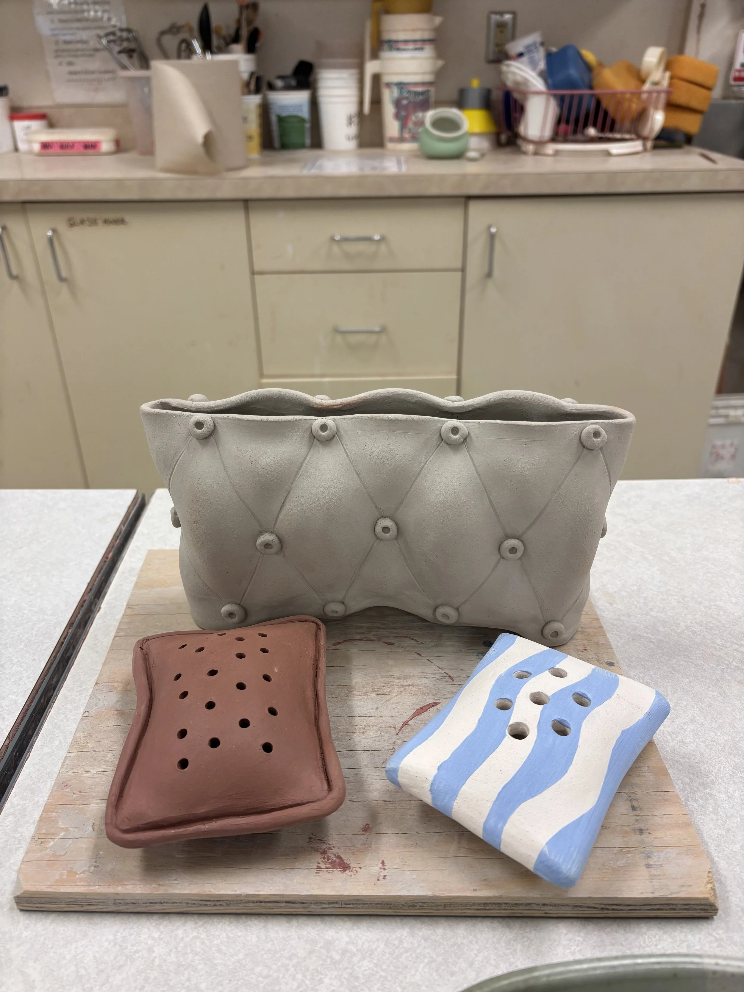 Unfired greenware ceramic tufted planter with scalloped rim alongside two handmade soap dishes — one terracotta with pierced holes, one with blue and white stripes — made by PVCC students in Malenka Luckett's class