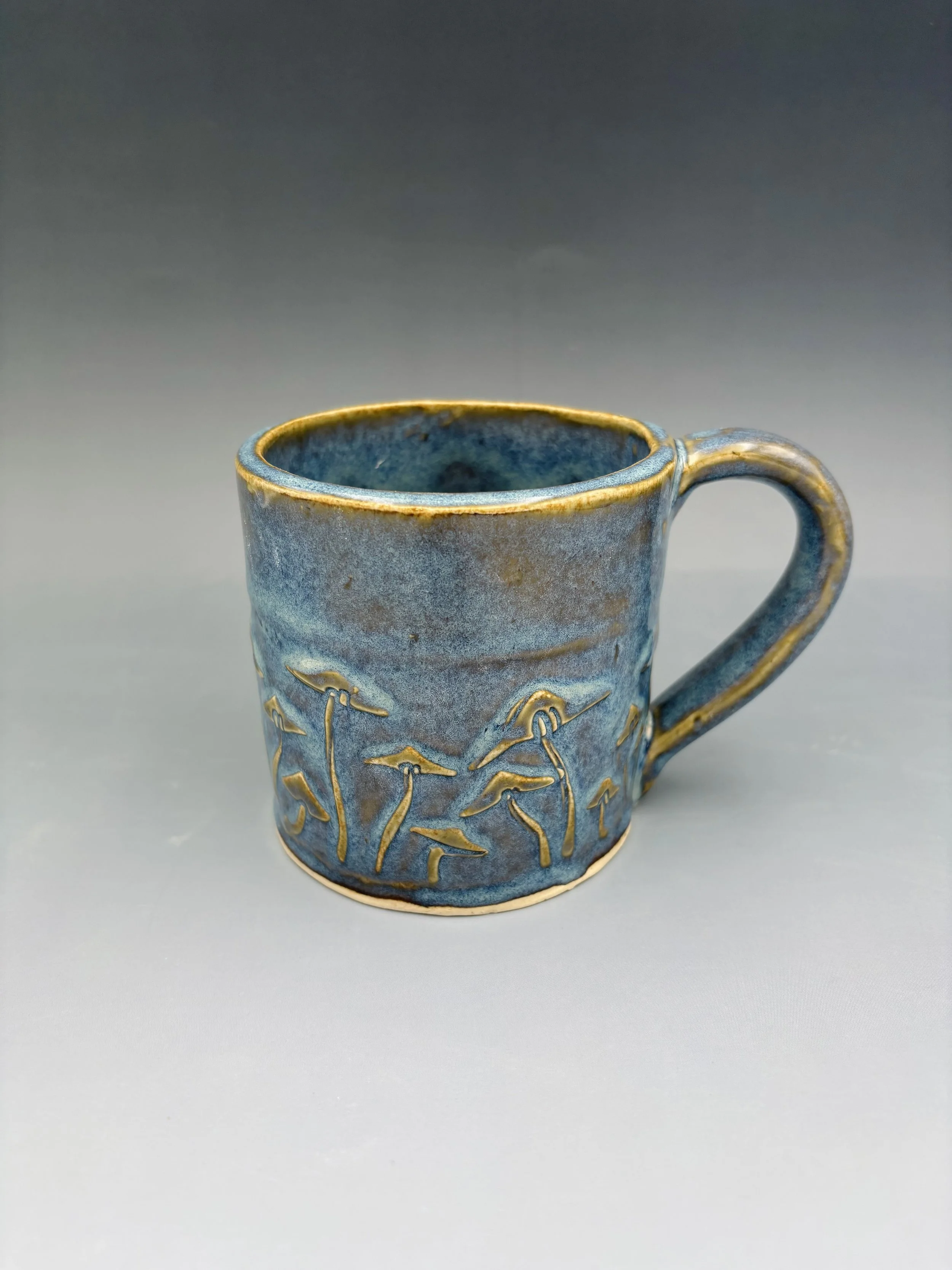 Handmade wheel-thrown ceramic mug in mottled blue and amber glaze with carved sgraffito mushroom illustration around the exterior, by student Reese McCardle made in Malenka Luckett's ceramics class