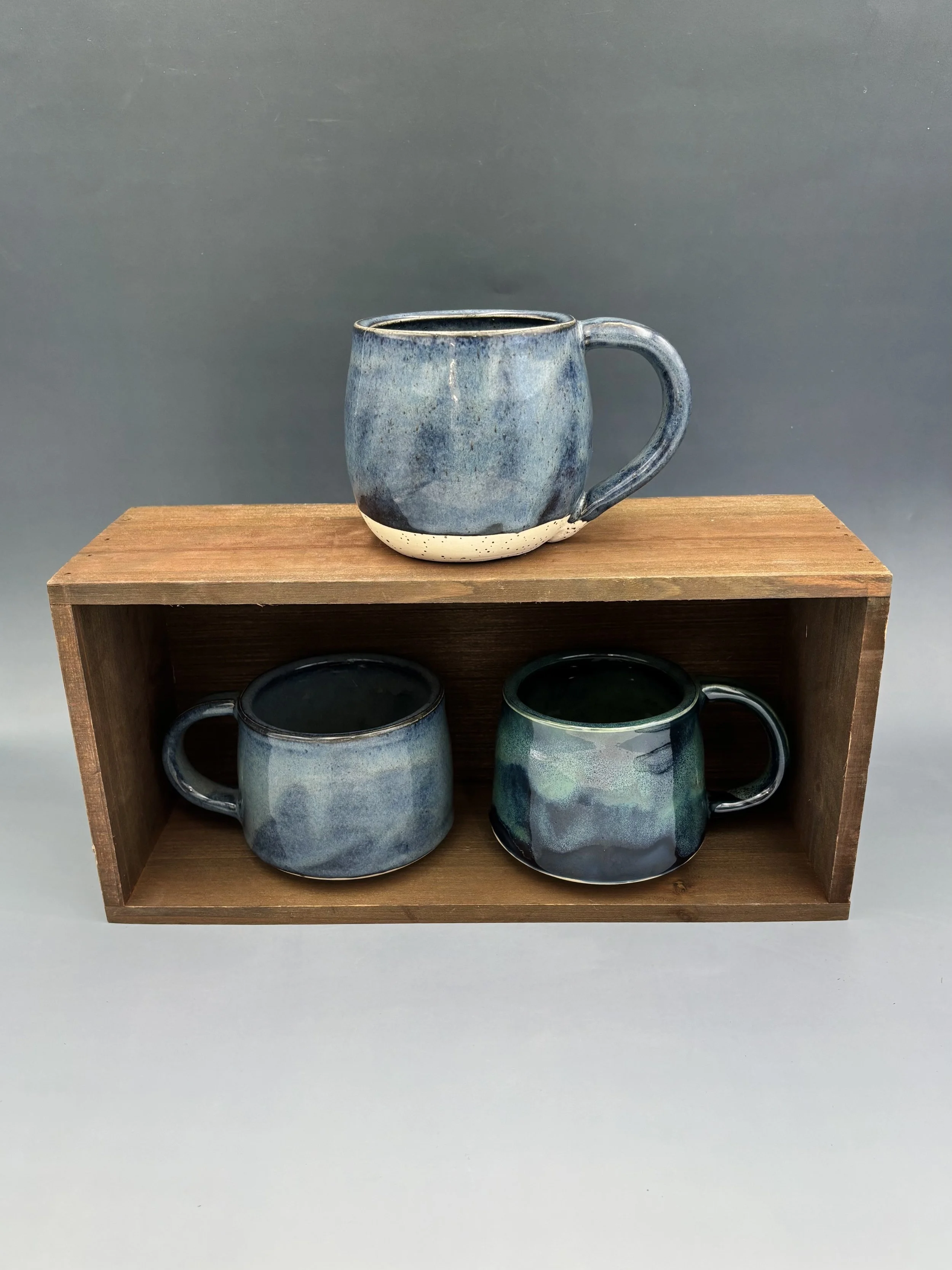 Three handmade wheel-thrown ceramic mugs by student Lisa Bowers in varying shades of deep blue and teal glaze, displayed on a wooden crate, made in Malenka Luckett's ceramics class