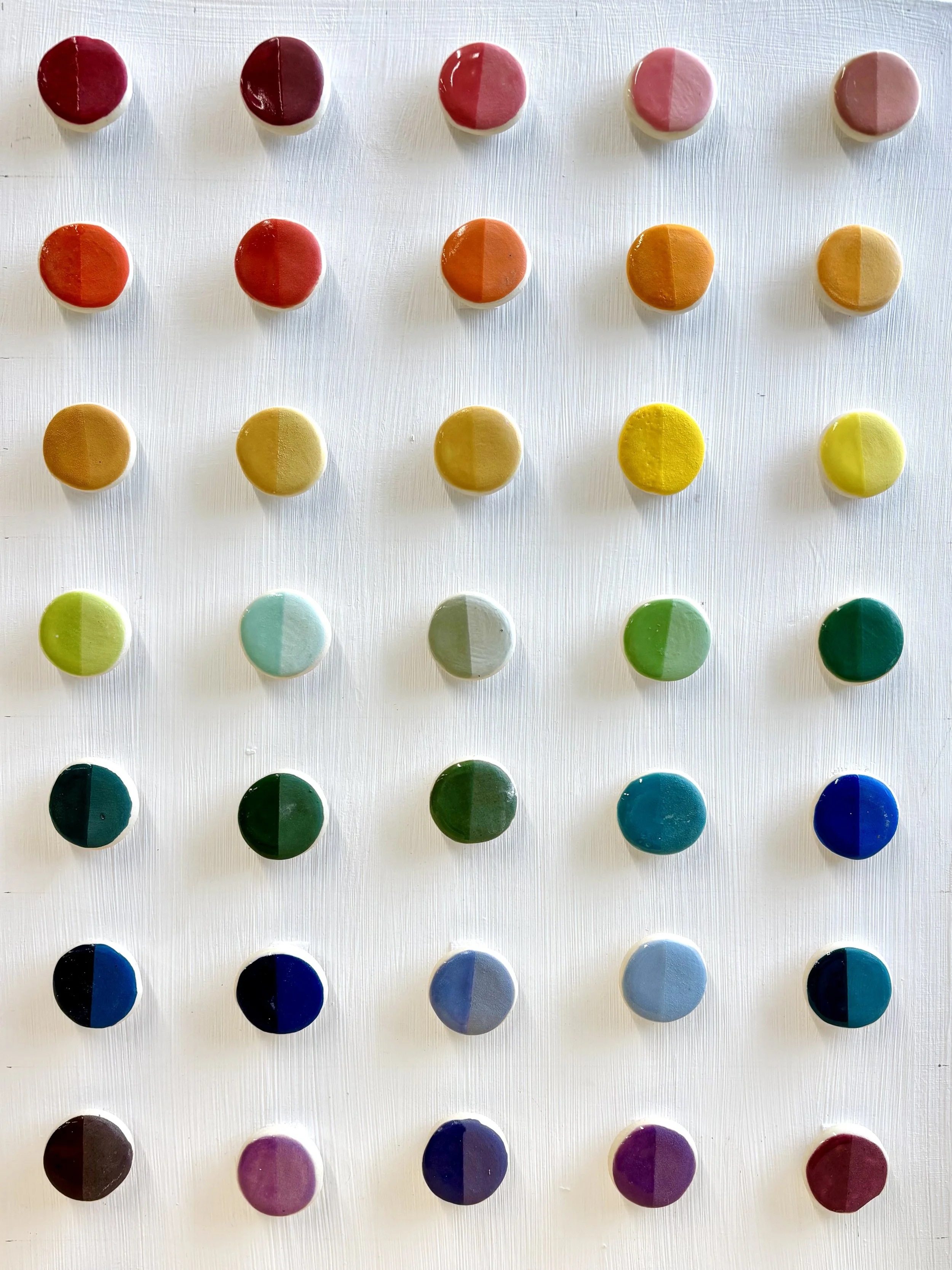 Grid of handmade ceramic underglaze test tiles arranged by color family, spanning the full spectrum from reds and oranges through yellows, greens, blues, and purples, used in Malenka Made's glaze development process