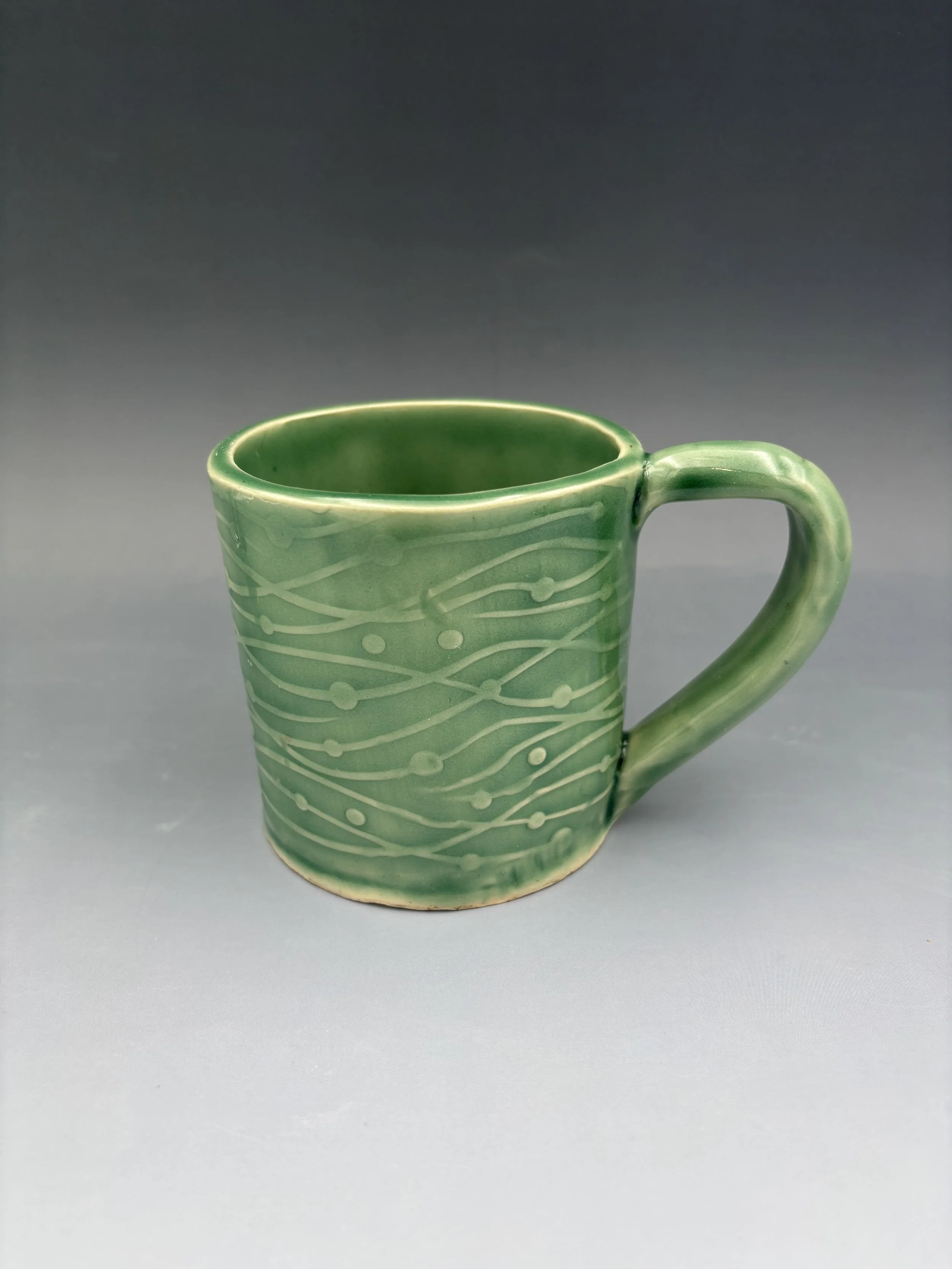 Handmade wheel-thrown ceramic mug in sage green glaze with carved sgraffito line and dot surface design, by student Reese McCardle made in Malenka Luckett's ceramics class
