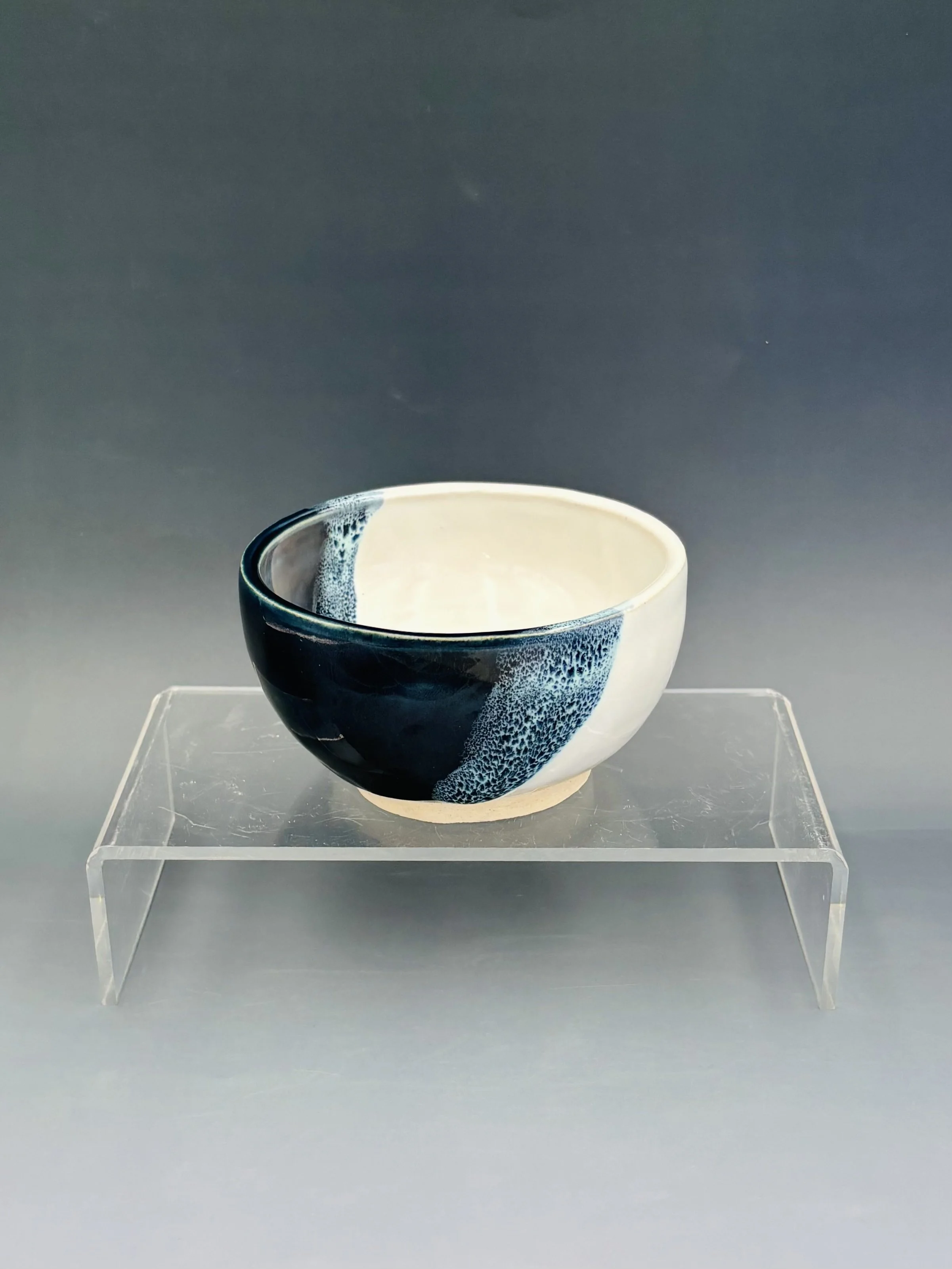 Handmade wheel-thrown ceramic bowl by student Lisa Bowers with bold navy blue glaze panels against a white glaze interior, displayed on a clear acrylic stand, made in Malenka Luckett's ceramics class