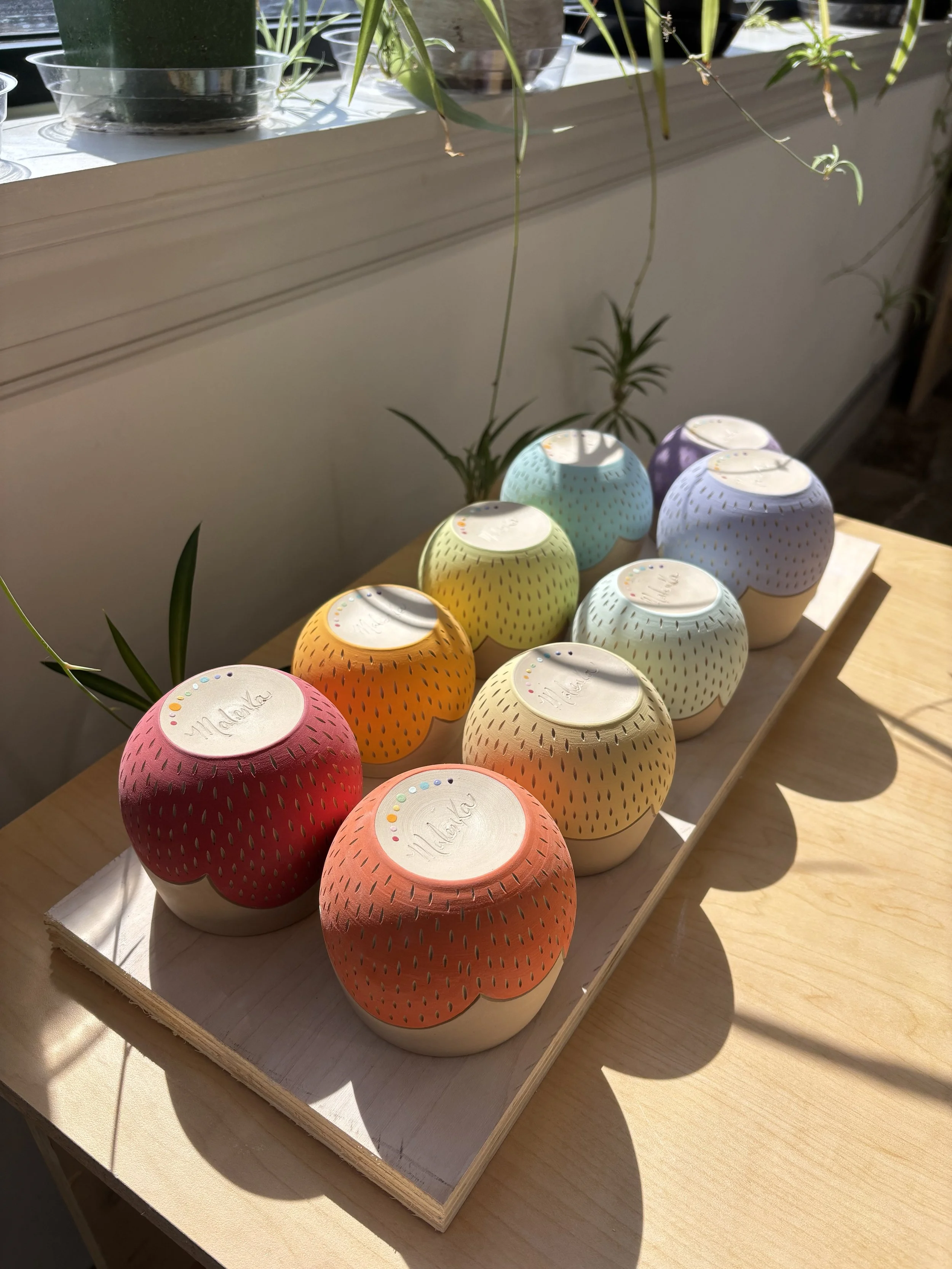 Eight handmade wheel-thrown ceramic tumblers in a full rainbow spectrum with scalloped edge design and hand-carved dot texture, signed by Malenka, displayed on a wooden board in natural light