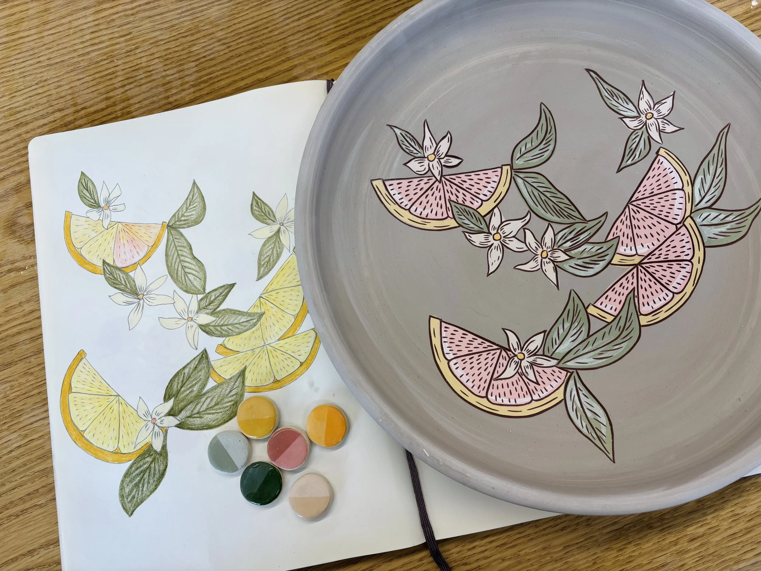 Handmade ceramic plate in progress with hand-illustrated grapefruit and citrus blossom design, shown beside a detailed color sketch and small underglaze test tiles