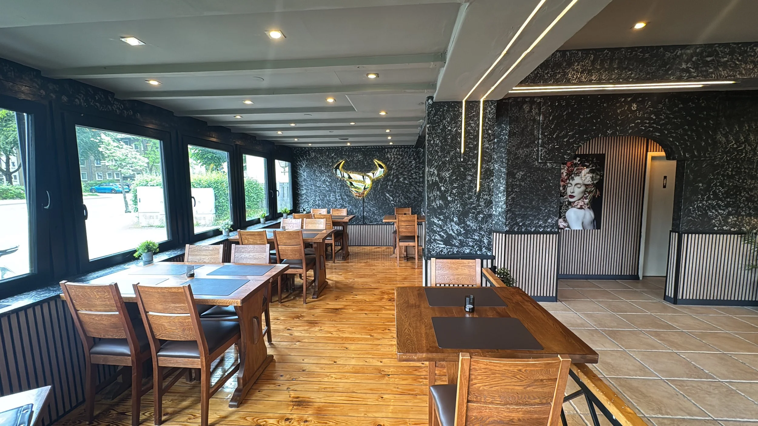 Innenansicht eines modernen Restaurants mit Holzfußboden, mehreren Holztischen und -stühlen, große Fenster mit Blick nach draußen, dunkle Wandgestaltung mit Kunstwerken und Beleuchtung.