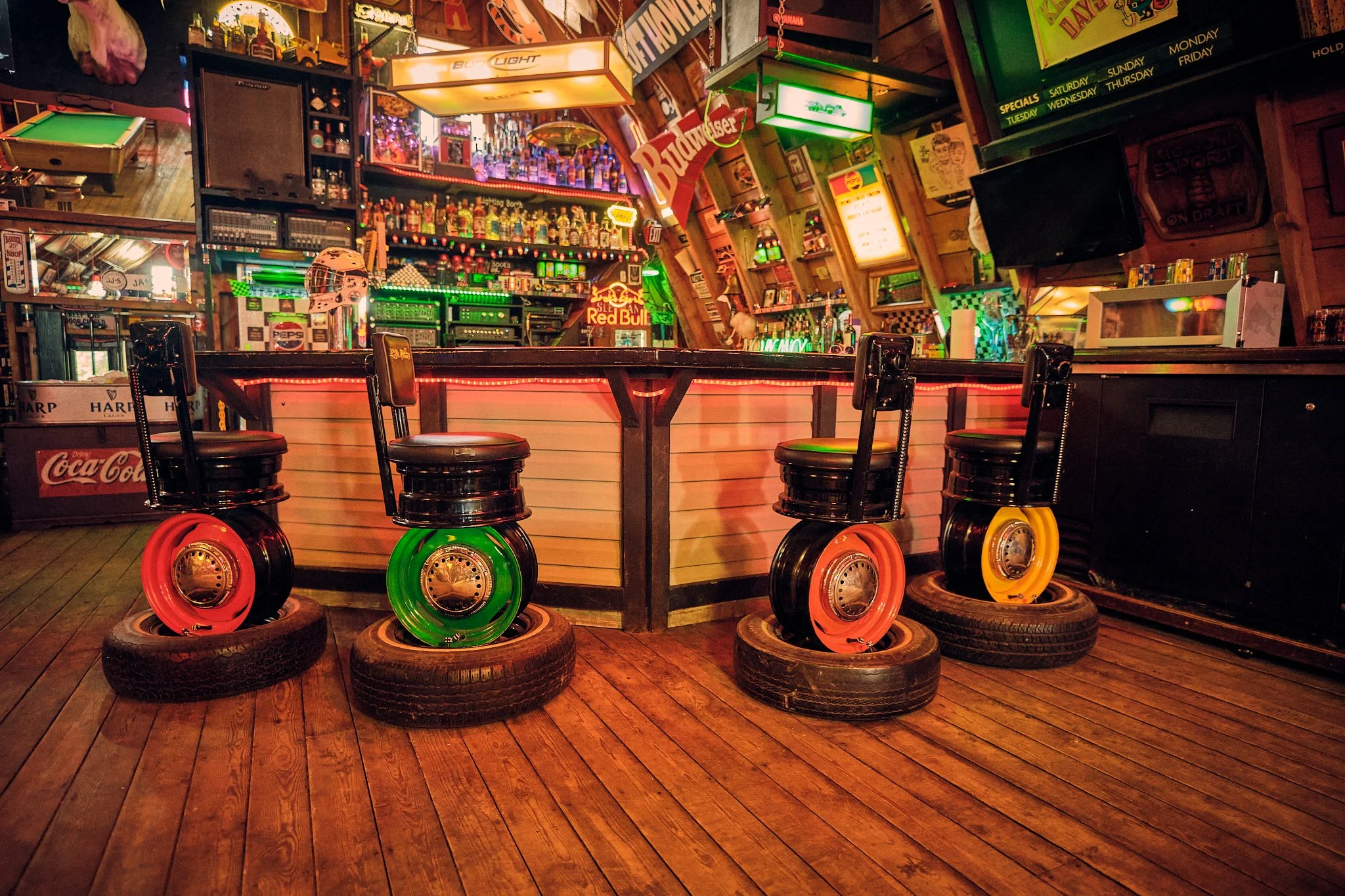 Inside a bar with a wooden counter and four colorful roller skate wheels placed on tires underneath. The background features neon signs, shelves with alcohol bottles, and various decorations including a dartboard and posters.