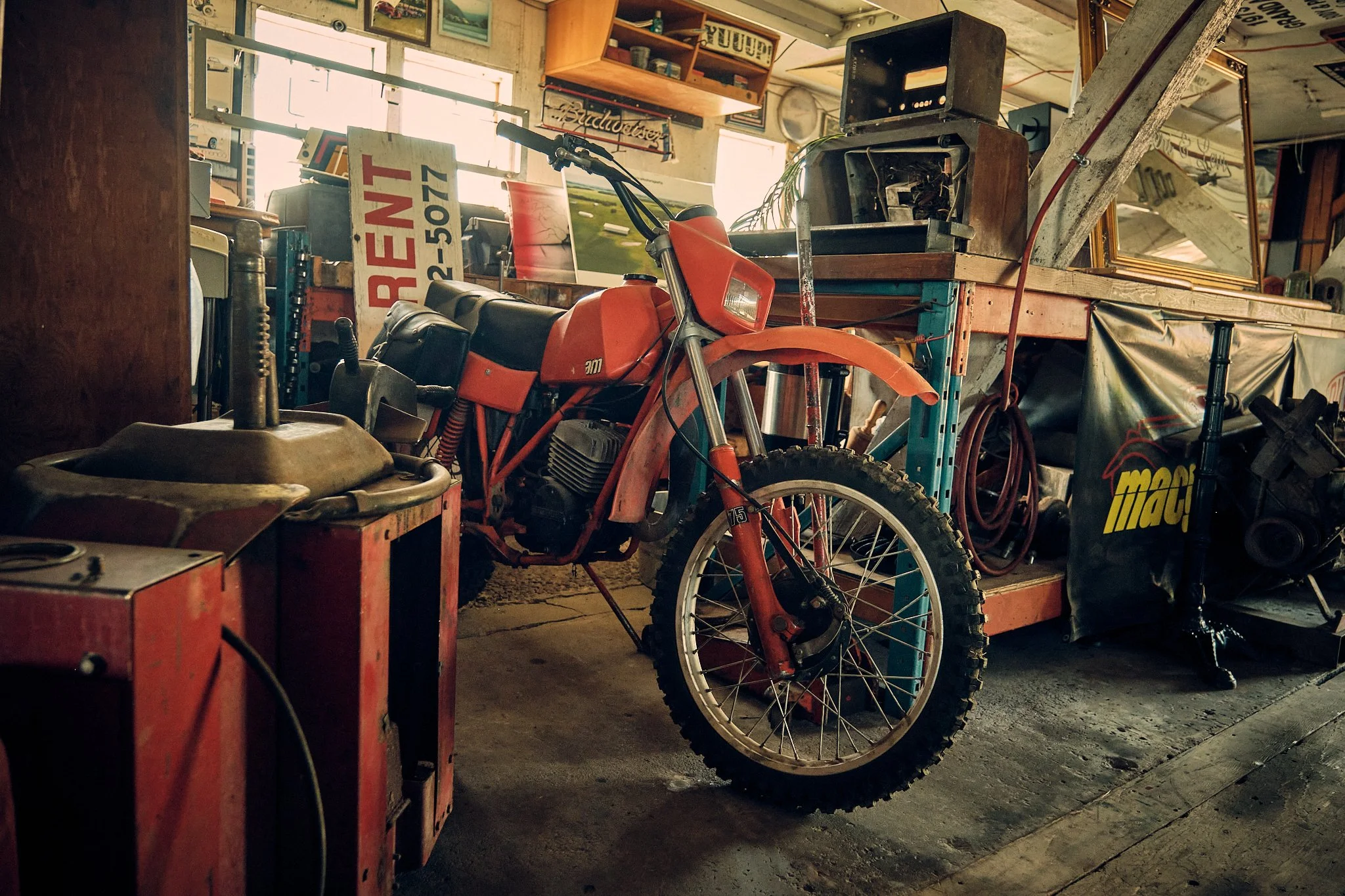 A cluttered vintage garage or workshop with a red dirt bike in the center, surrounded by various tools, signs, and old furniture, including a large window and framed pictures on the wall.