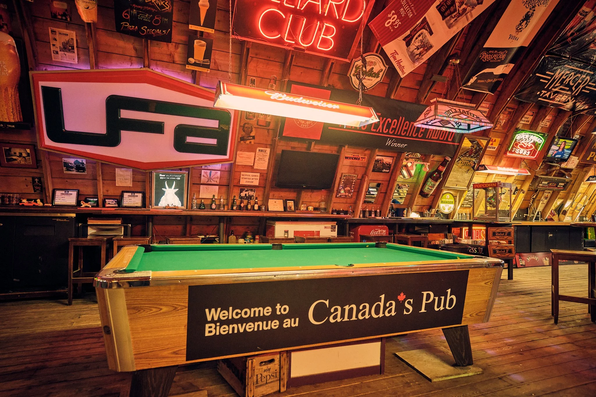 Interior of a Canadian pub with wooden walls decorated with neon signs and posters, a pool table in the foreground, and a table with a sign welcoming guests to Canada.