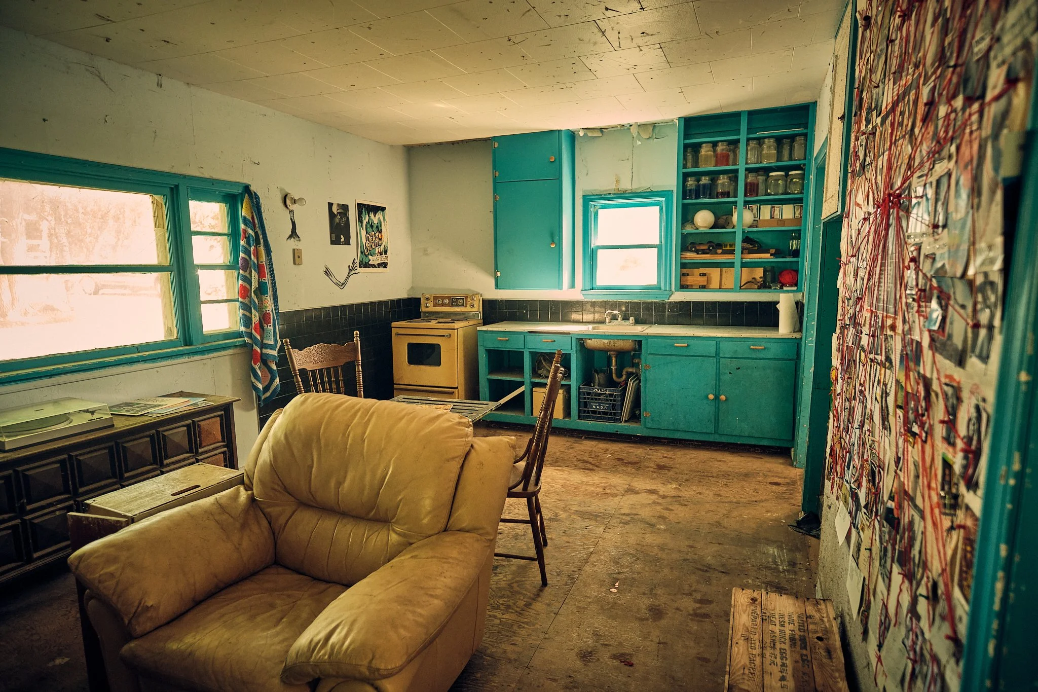A cluttered kitchen with teal cabinets, yellow stove, and two windows, with a beige recliner and a dark wooden dresser nearby, and various pictures on the walls.