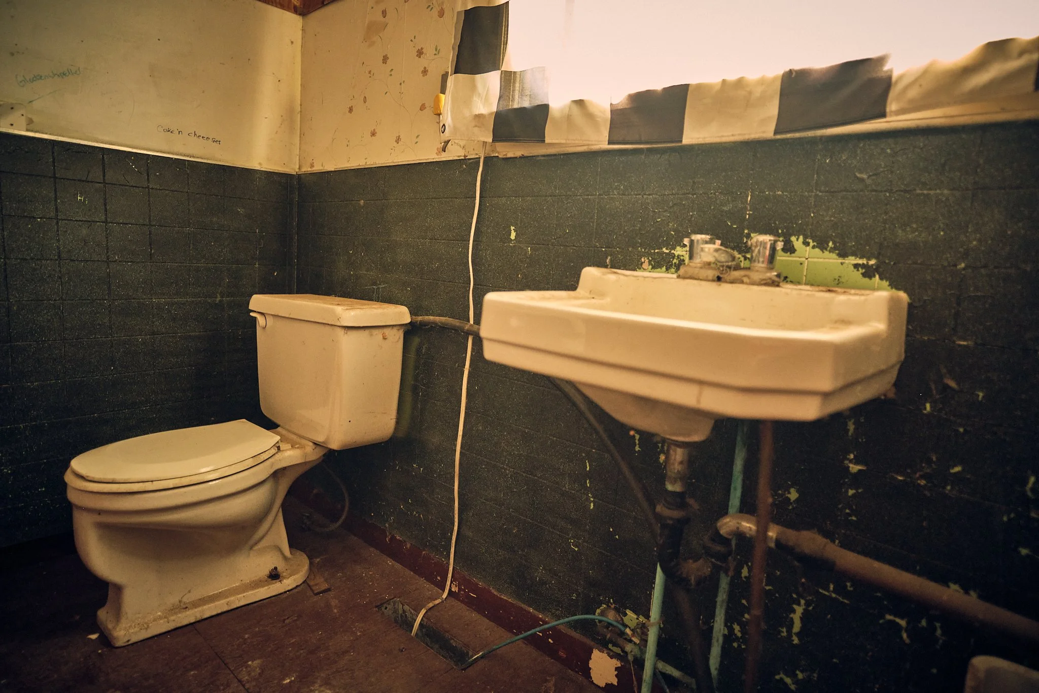 A dirty, grimy bathroom with a white toilet, an old sink, and peeling wallpaper. The room appears neglected and stained.