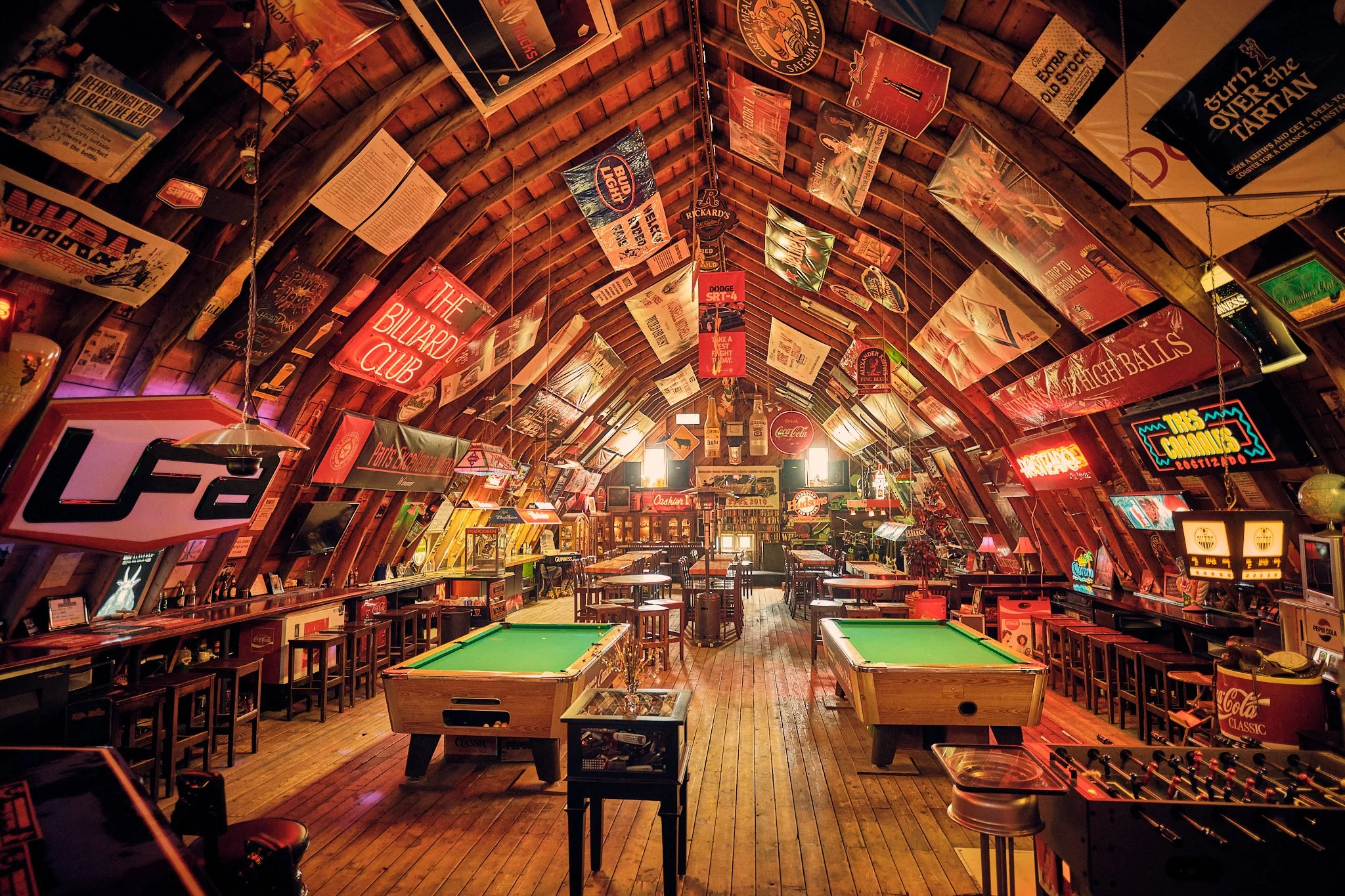Interior of a bar or pub with wooden ceilings and walls decorated with numerous vintage signs and posters, pool tables at the center, and tables and chairs along the sides.
