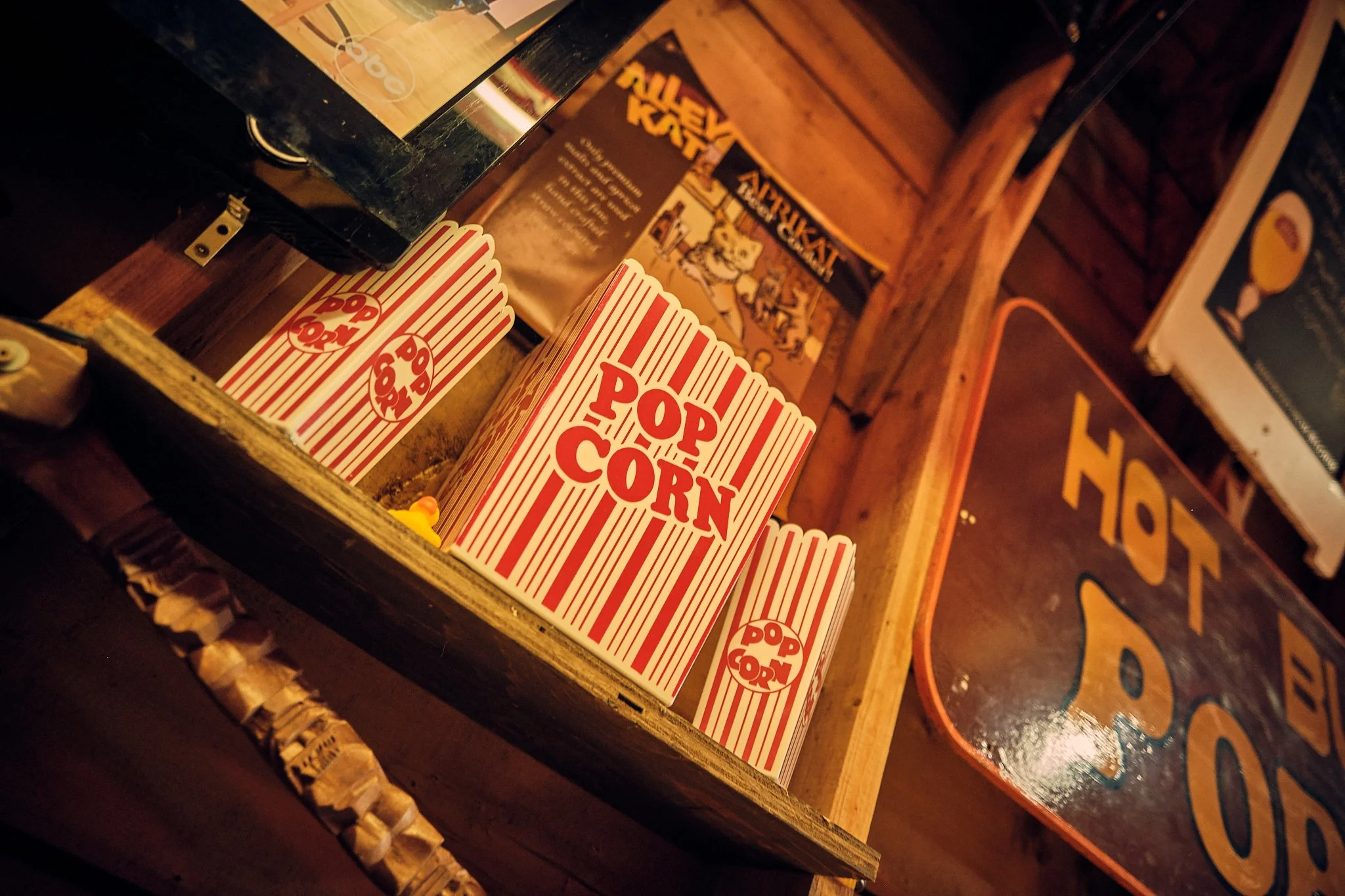 Close-up of stacked popcorn containers with red and white stripes on a wooden surface, background includes a partially visible menu board and a retro-style sign, in a dimly lit setting.