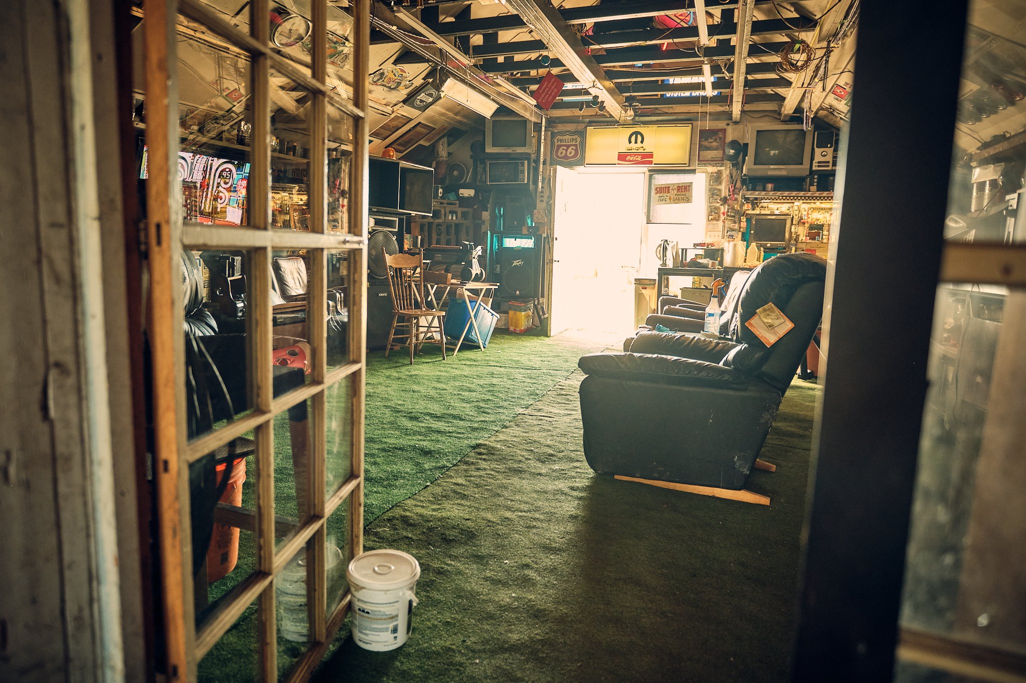Interior of a cluttered casual room or garage with vintage signs, television, chairs, and various items, illuminated by natural light from an open door.