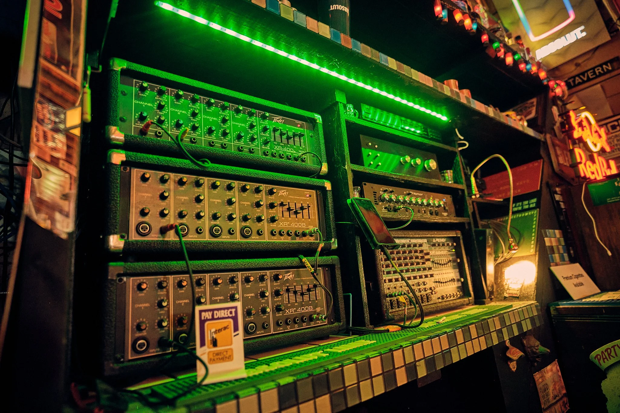 A collection of music equipment including various audio synthesizers, mixers, and control panels illuminated by green lighting in a dimly lit environment.