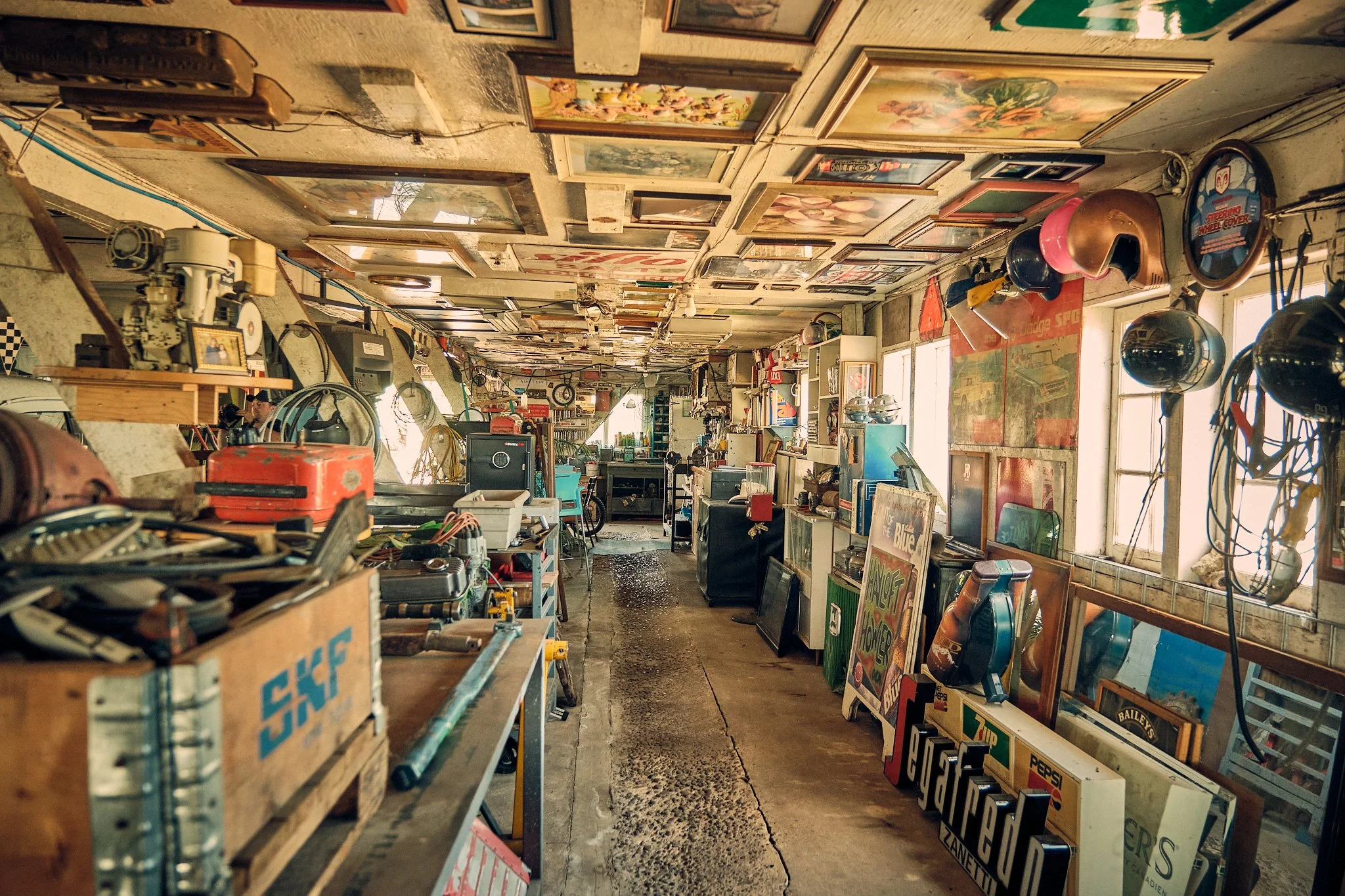 A cluttered vintage store or flea market with numerous framed pictures and posters hanging on the walls, shelves filled with assorted items, helmets hanging near the window, and various signs and objects scattered around inside.