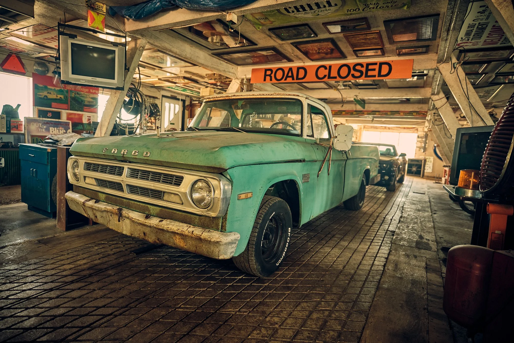 A vintage green Fargo pickup truck parked inside a cluttered garage with a "Road Closed" sign hanging from the ceiling. The garage contains various items including an old television, framed pictures, and other auto memorabilia.