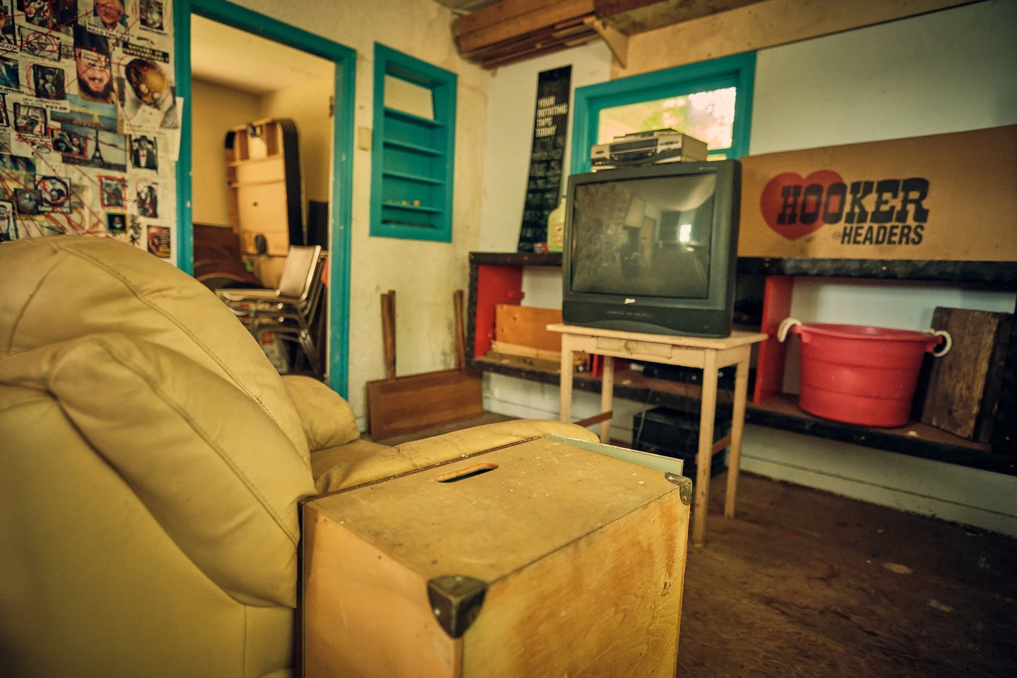 A cozy living room with a tan leather sofa, a wooden coffee table, a vintage TV, and a red bucket. The room has a wall with a collage of photos and a poster, and a blue-framed window with a view outside. The ceiling has exposed wooden beams, and ther