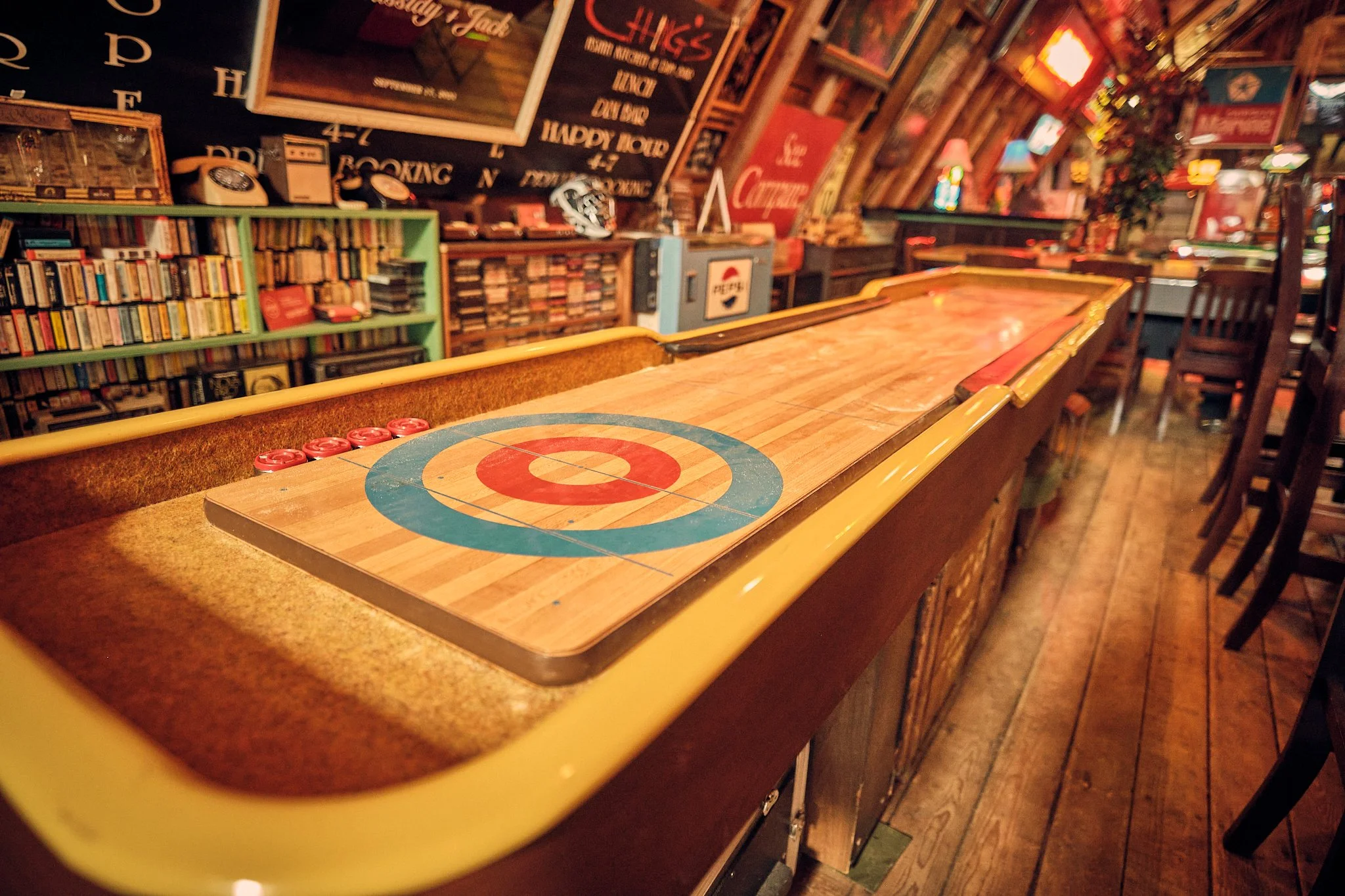 Interior of a cozy bar or game room with a shuffleboard table in the foreground, shelves filled with books, vintage telephones, and decor, with colorful signs and dim lighting creating a warm, inviting atmosphere.