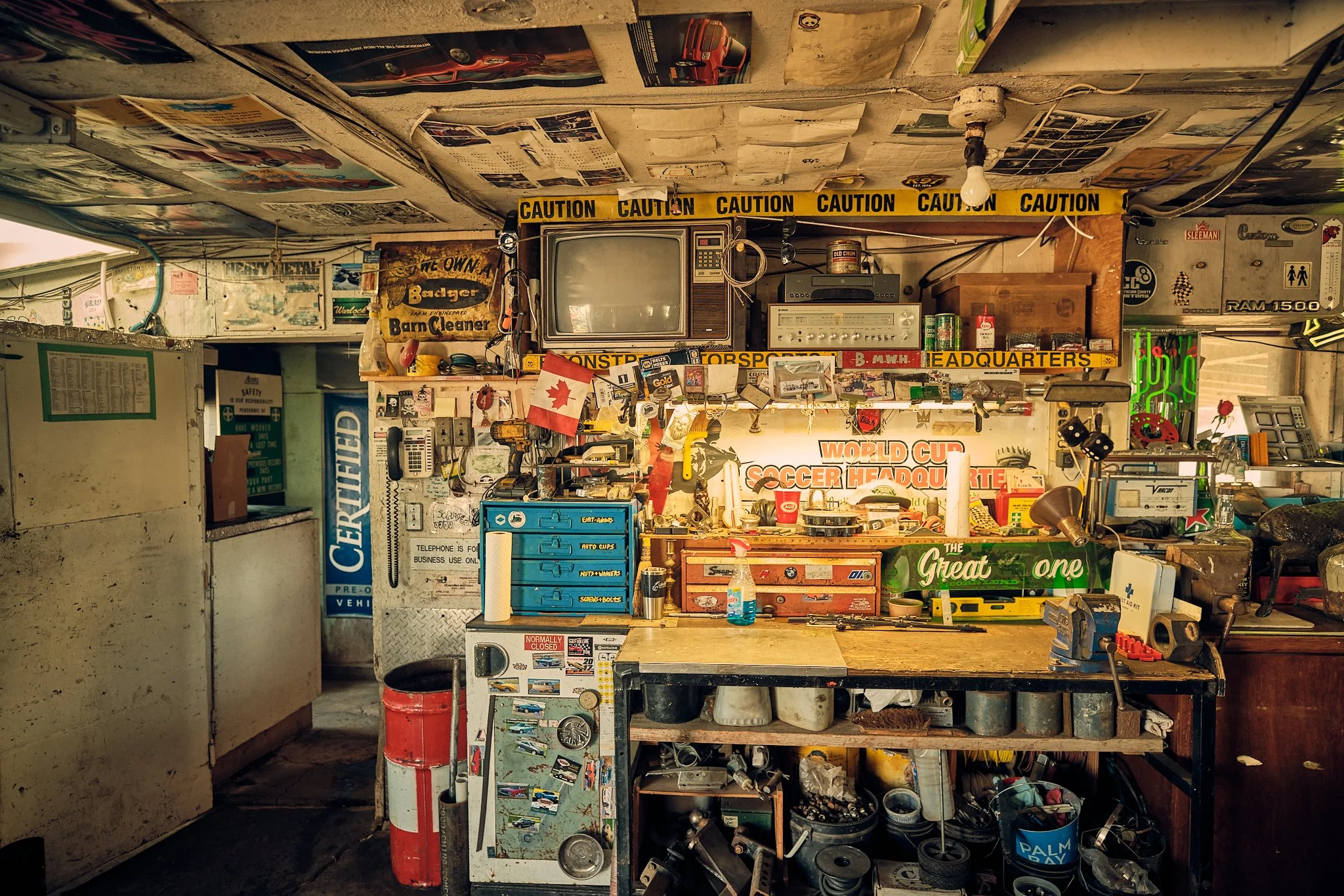 A cluttered garage workbench filled with various tools, posters, a vintage television, a stereo, and numerous small items and stickers on the wall and shelves.