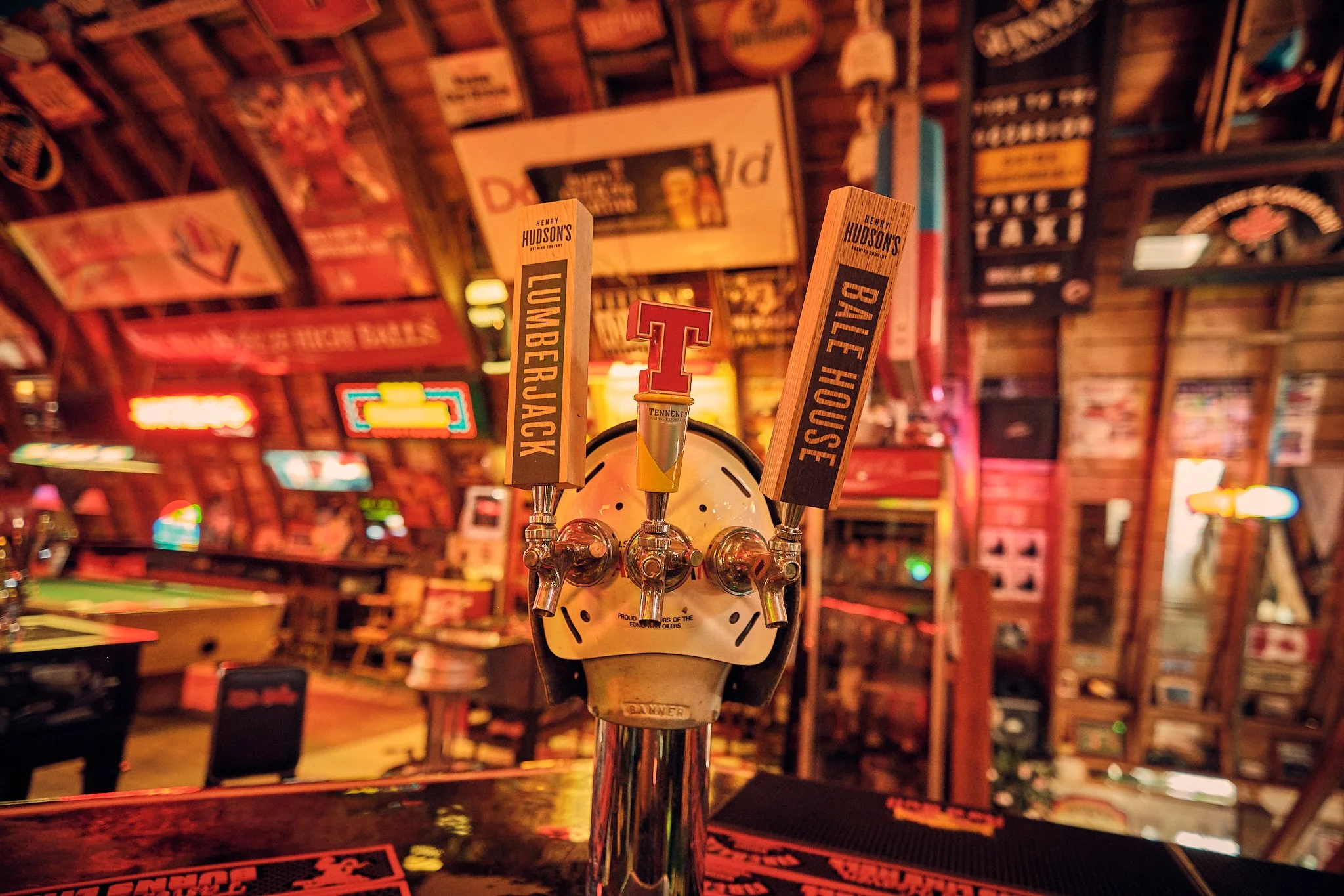 Close-up of four beer taps labeled 'Lumberjack,' 'Bale Fiance,' 'Tennessee,' and another without visible label in a bar with wooden decor and colorful signs.