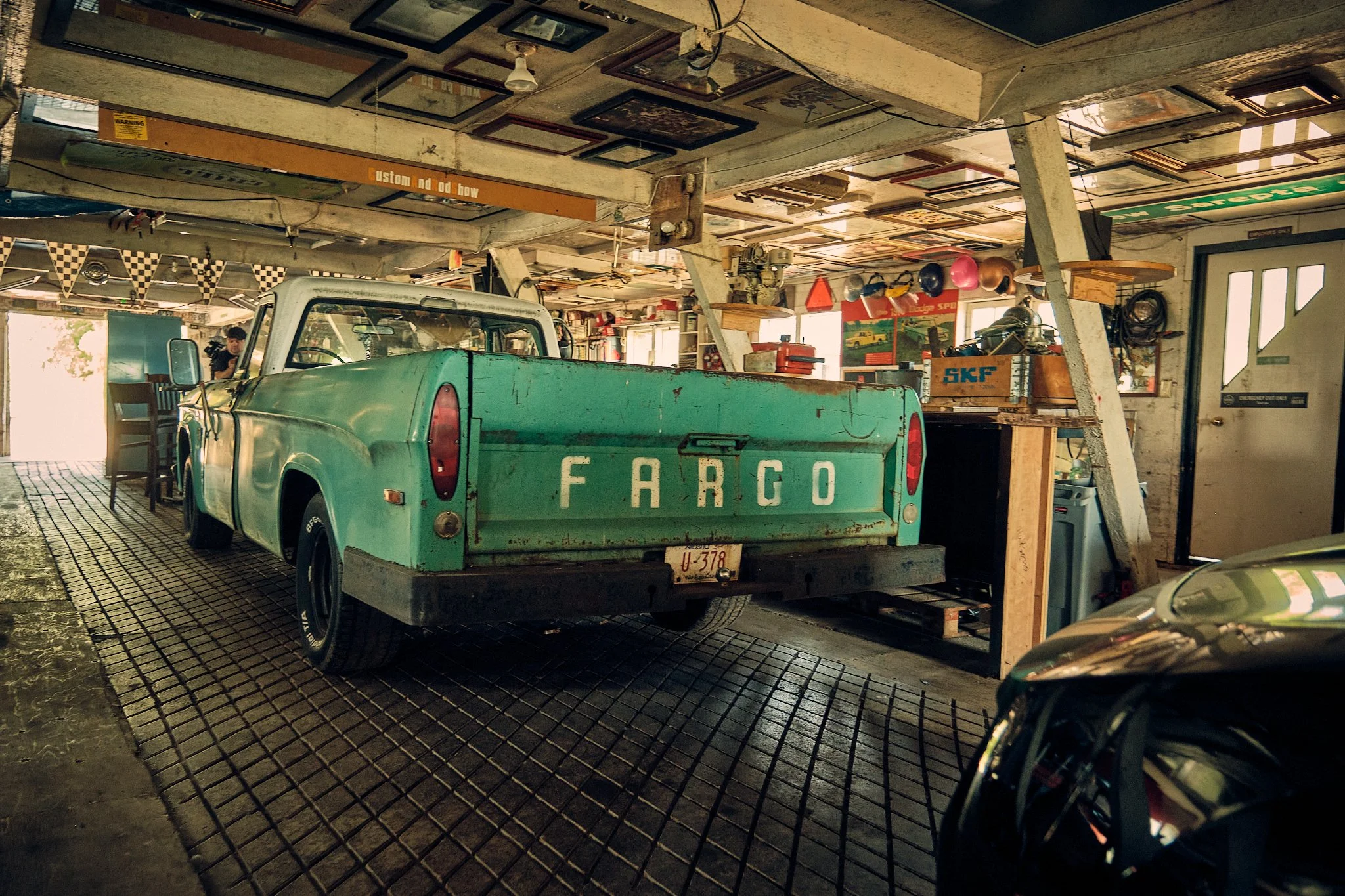 Inside a vintage garage with an old green Ford truck, various tools, signs, and framed pictures hanging on the ceiling and walls.