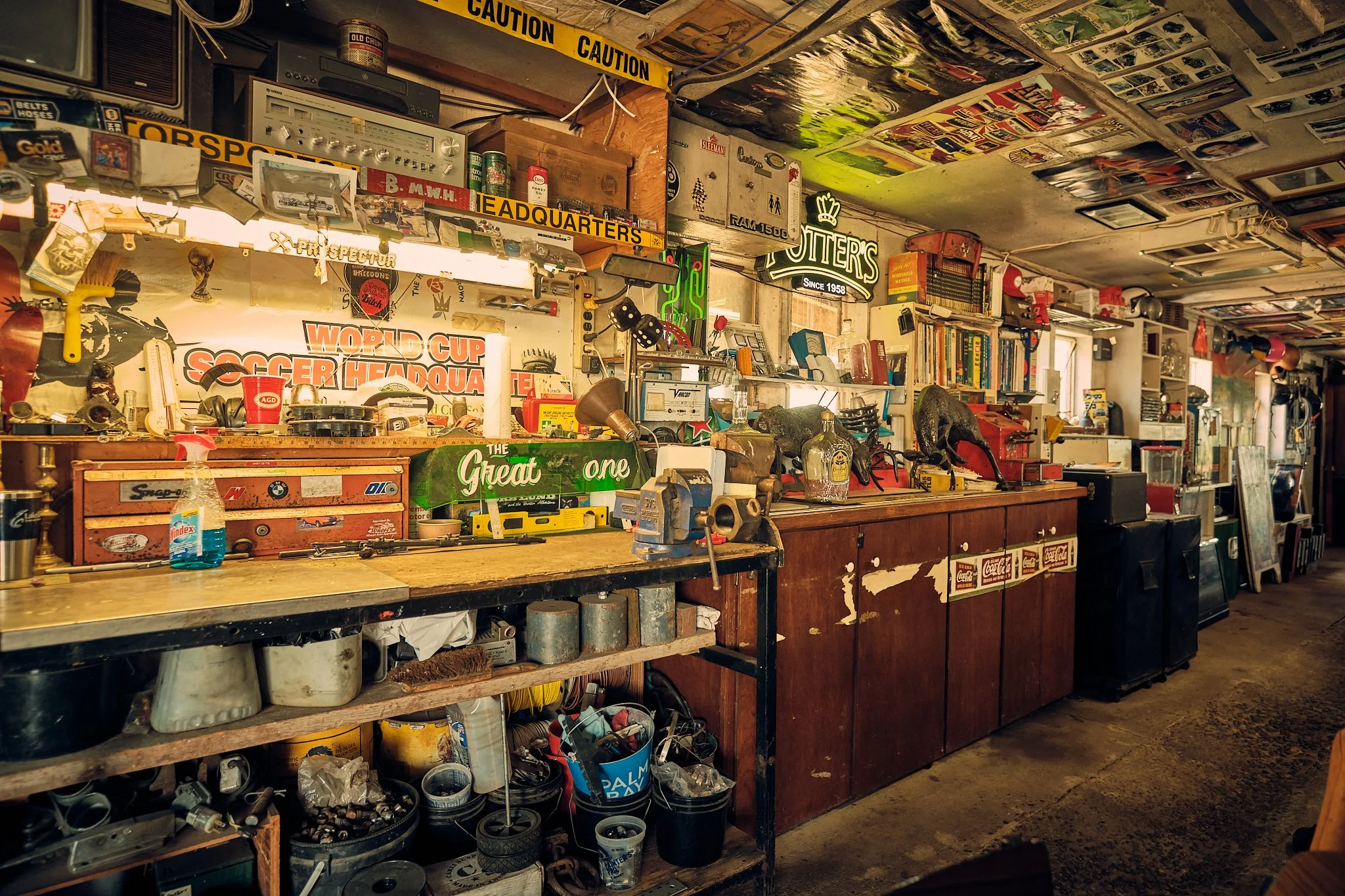 An interior view of a cluttered workshop or garage filled with various tools, collectibles, books, and memorabilia, including vintage signs, a stereo system, and shelves stocked with items, with caution tape hanging across the ceiling.