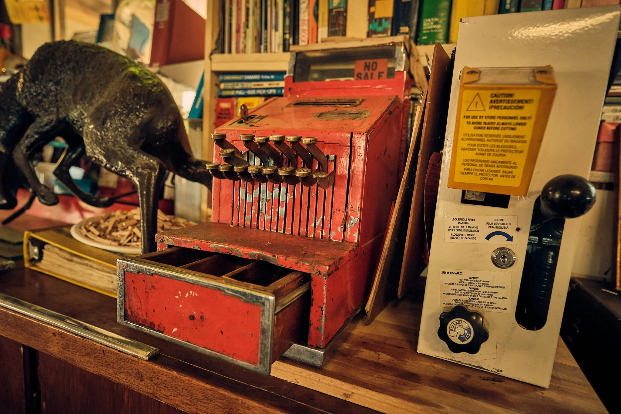 A vintage red coin-operated capsule toy vending machine with a rusted exterior, placed on a wooden surface. To its right, there is a white panel with a yellow warning label. In the background, there are shelves filled with books.